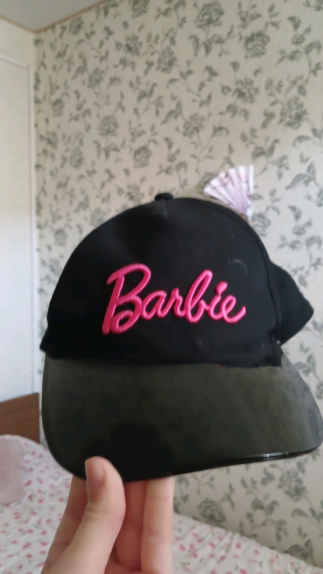 A black baseball cap with "Barbie" embroidered in pink cursive lettering is held in front of floral wallpaper. The brim of the cap appears to be dark green. A portion of a bed with floral bedding is visible in the background. The image is taken indoors. The location is not specified in the image or context.