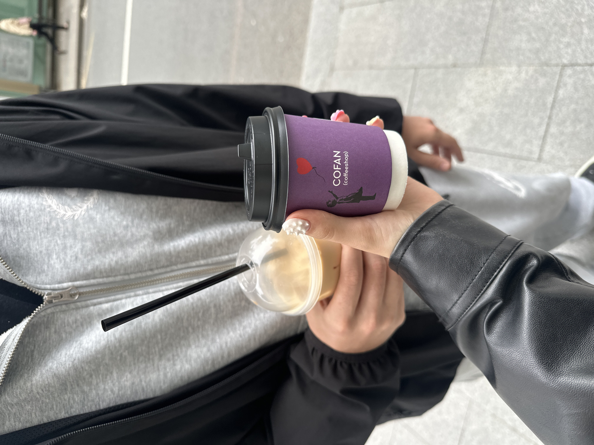 The image shows a close-up of two people holding coffee cups. One person, wearing a black leather jacket and a grey zip-up hoodie, holds a clear plastic cup with a light brown beverage and a black straw. The other person, wearing a black jacket and grey sweatpants, holds a purple paper coffee cup with a black lid. The purple cup has a logo that reads "COFAN (coffeeshop)" and features a silhouette of a person holding a red heart balloon. The background suggests an urban environment with a paved sidewalk and blurred figures in the distance, potentially indicating a street in Moscow, Russia. The lighting is bright, suggesting daylight.