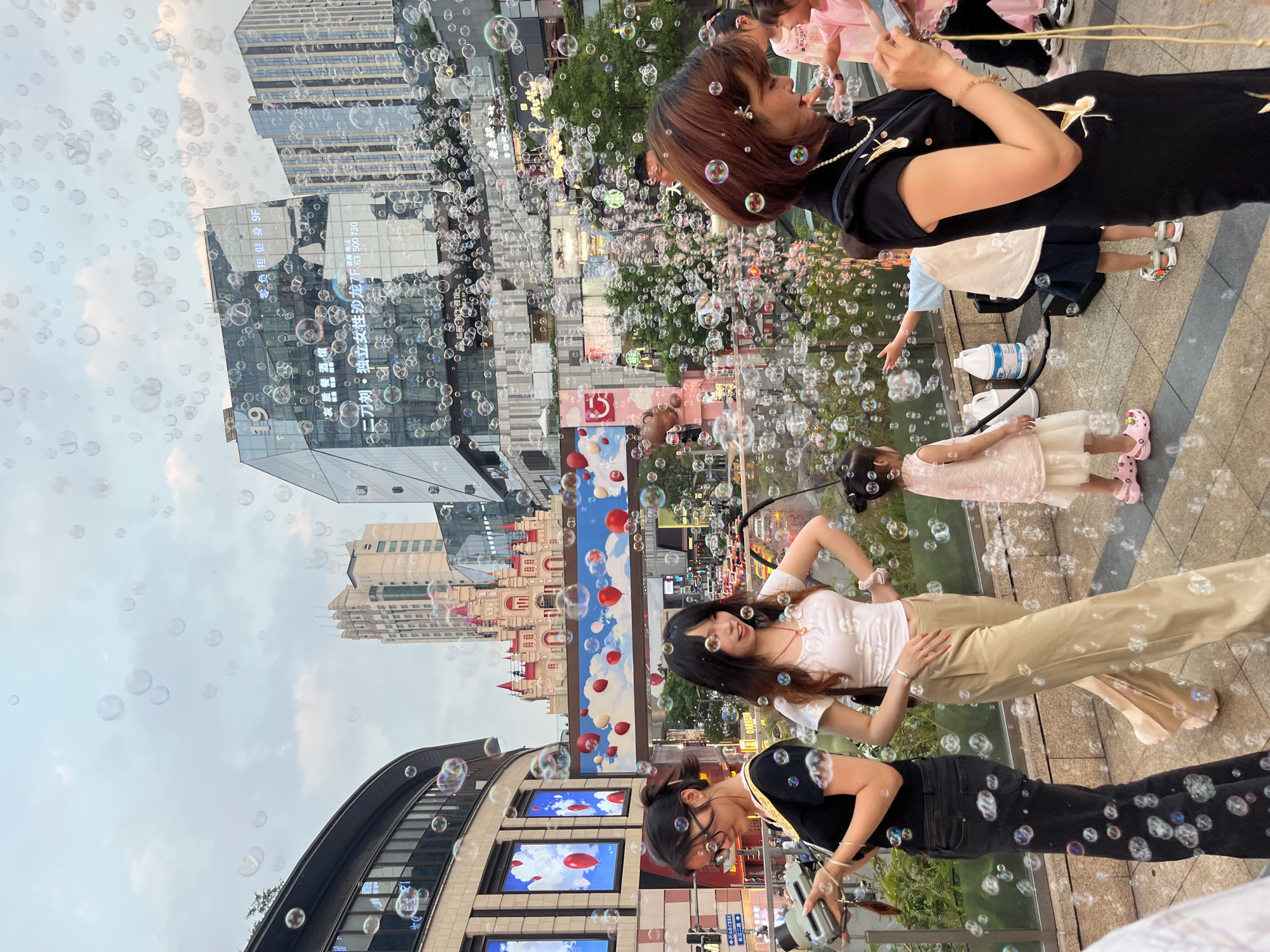 The image captures a vibrant urban pedestrian area in Yuzhong District, China, densely populated with floating soap bubbles under a lightly overcast sky.
In the foreground, on the right, a woman with reddish-brown hair, adorned with a pearl necklace and wearing a black short-sleeved cheongsam dress featuring gold crane embroidery, looks down at a smartphone held in her left hand. To her immediate left, a young child in a pink dress and pink Crocs shoes observes the scene. Central to the mid-ground, a black bubble machine operates on the paved ground, actively generating the numerous bubbles; two large white plastic containers, likely holding bubble solution, sit beside it. A child in a white top and dark shorts stands near the machine, extending an arm towards the bubbles. Further left, a young woman with long dark hair, dressed in a white short-sleeved top and light brown wide-leg trousers, stands with her right hand on her hip, gazing towards the right. Adjacent to her, a woman with dark hair, wearing glasses, a black short-sleeved top, and dark jeans, holds a grey instant camera, looking down at it.
The background reveals a bustling cityscape. On the left, a multi-story building with a curved glass facade displays large screens featuring images of red balloons against a blue sky. In the center, a prominent red and white castle-like structure stands atop a building, connected by a pedestrian bridge decorated with red balloon motifs. To the right, several modern glass-clad high-rise buildings are visible, displaying illuminated Chinese commercial signage, including "独立女性沙龙 7F" (Independent Women's Salon 7F) on one facade. Distant street traffic and additional high-rise structures are discernible beneath elevated pedestrian pathways. The sky is a pale blue-grey with scattered clouds. The ground is paved with light-colored tiles, and sparse greenery is visible along the urban architecture.