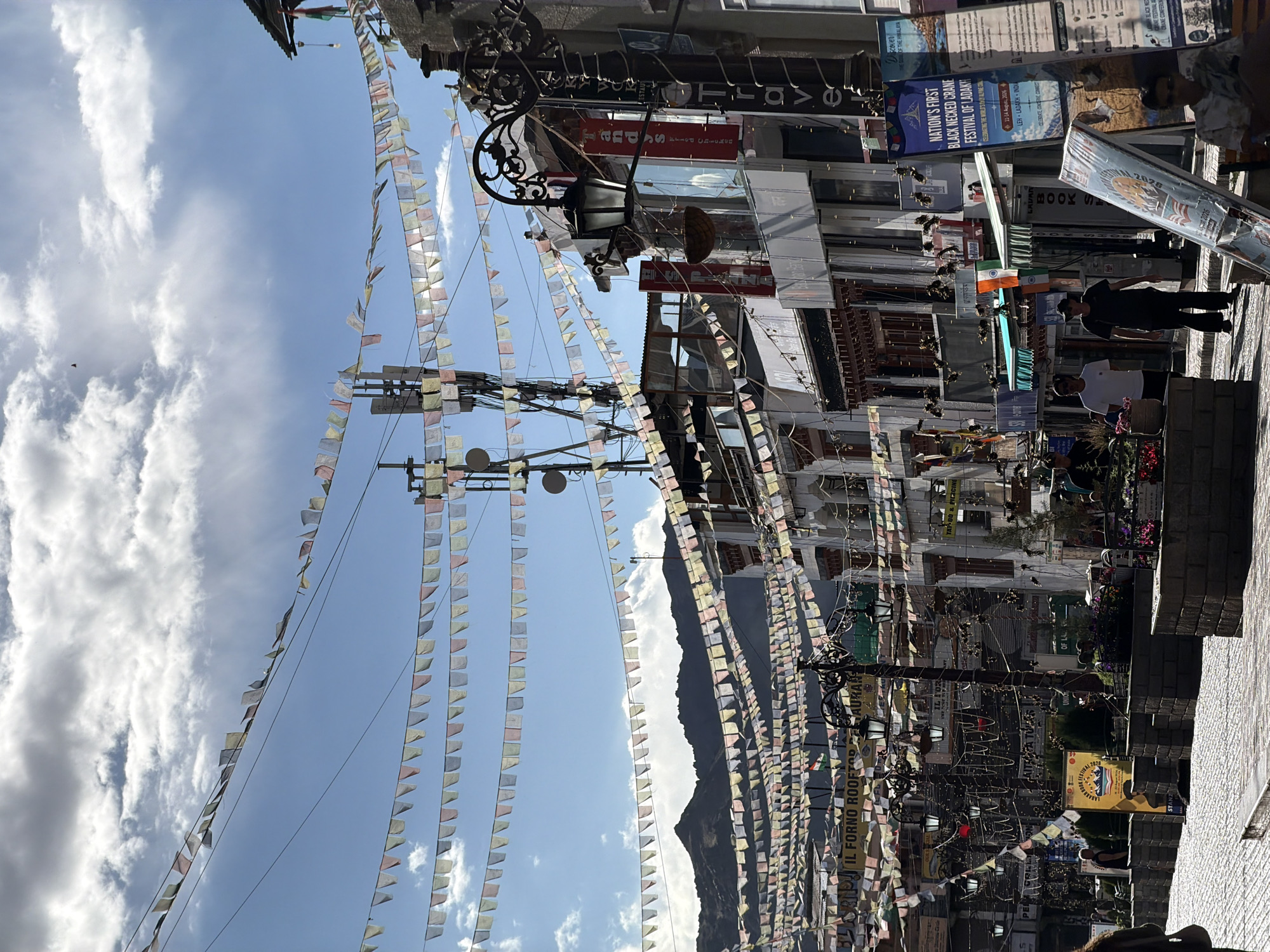 This image depicts a street scene in the city of Leh, India, likely on a bright, sunny day with scattered clouds. The foreground shows a cobblestone street lined with buildings on the right. Several strings of colorful prayer flags are strung across the street, creating a festive atmosphere. The buildings appear to be commercial establishments, with signage visible, including "IL FORNO ROOFTOP RESTAURANT" and "TRAVEL." There are streetlights and utility poles with antennas.
In the middle ground, people are seen walking and standing. Two men are walking on the street, one in a white t-shirt and the other in a dark t-shirt and hat. Another person is seated in the lower right corner. Further into the background, more people are visible, and a large banner advertising the "NATION'S FIRST BLACK NECKED CRANE FESTIVAL OF LADAKH" is prominently displayed. An Indian flag is also visible flying from a building.
The background features a range of mountains, characteristic of the Himalayan region where Leh is located. The sky is a bright blue with white clouds, and the overall lighting suggests it is daytime. The scene appears to be a busy commercial area, likely a popular tourist spot given the signage and the festive decorations.