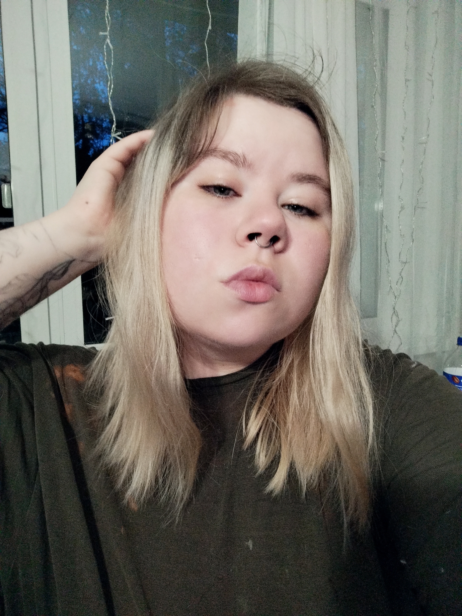 A close-up selfie captures a person with fair skin, blonde hair, and a nose ring. The person is wearing a dark green shirt. The person's left hand is raised, touching their hair. The background features a window with a view of a dark, possibly nighttime, scene outside. There are decorative string lights hanging in front of the window. A sheer curtain partially obscures the view on the right side. The location is Unknown, Unknown.