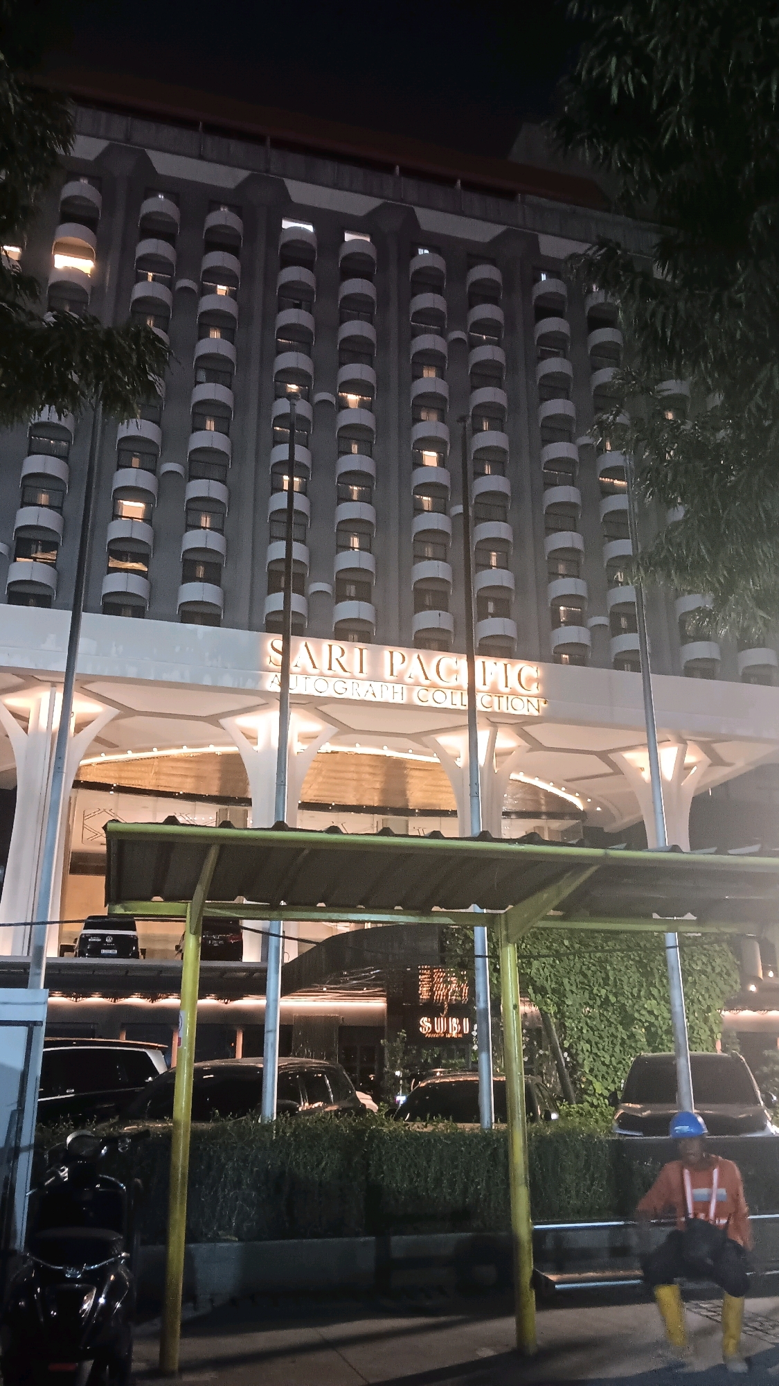 The image captures the exterior of the Sari Pacific Hotel, an Autograph Collection hotel in Central Jakarta, Indonesia, at night. The hotel's facade is characterized by repeating rows of balconies, illuminated from within, creating a rhythmic pattern against the dark sky. The entrance area is lit with warm, diffused lighting, highlighting the architectural details of the canopy and support columns. In the foreground, a covered waiting area with yellow and green accents is visible, sheltering parked cars and a parked motorcycle. A construction worker, wearing a blue hard hat, an orange long-sleeved shirt, dark pants, and yellow boots, is seated on a bench. The overall scene is one of urban nighttime ambiance, with a focus on the hotel's architecture and street-level activity.