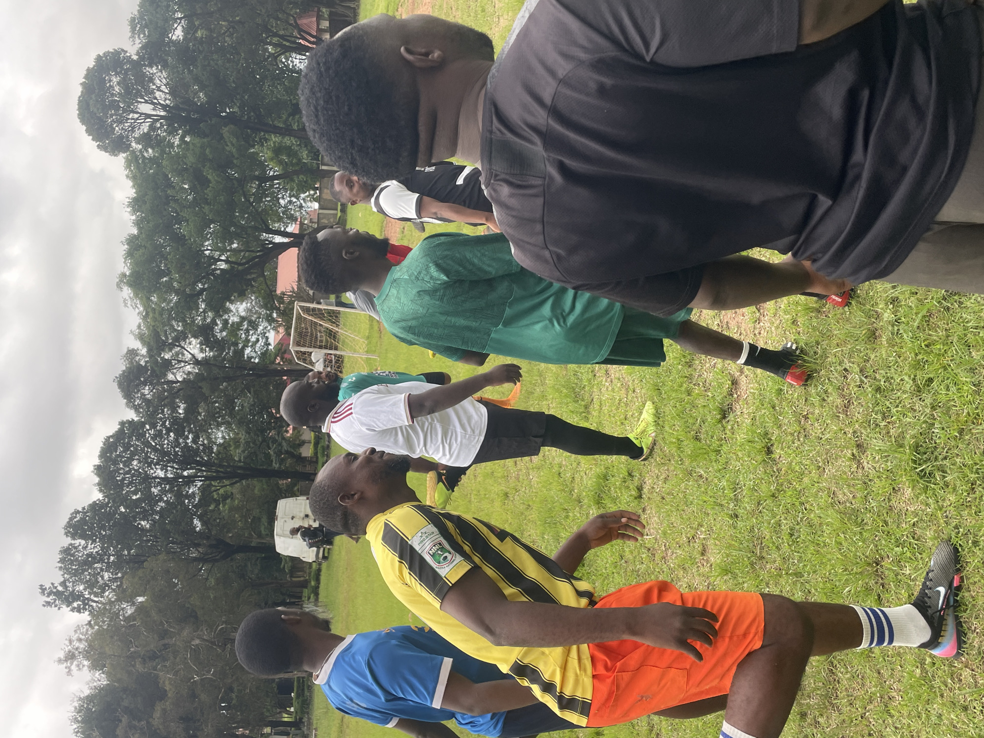 This image depicts a group of men gathered on a grassy field, possibly for a sports activity. The environment appears to be outdoors with trees and a cloudy sky in the background. Several individuals are wearing athletic attire, including jerseys and shorts. In the distance, a white van and a soccer goal are visible. One man in the foreground is wearing a yellow and black striped jersey with orange shorts and blue-striped socks. Another man to his left wears a blue jersey. Further back, men in green and white jerseys are also present. The overall impression is that of a casual sporting event or practice session taking place in an open area. The location is identified as Jos, Nigeria, suggesting an urban or semi-urban setting.