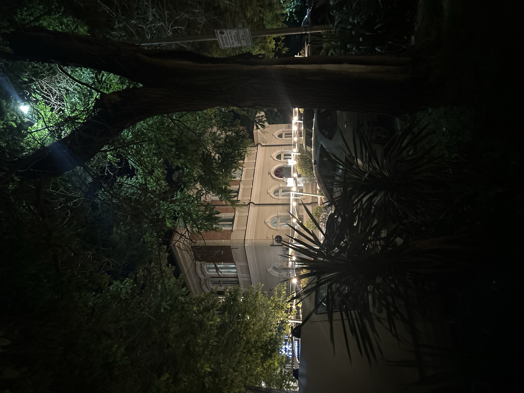 At night, a three-story building is viewed through trees and bushes in Burgas, Bulgaria. The building has arched windows. Strings of lights decorate the front of the building. Parked vehicles are in front. The image is dark.