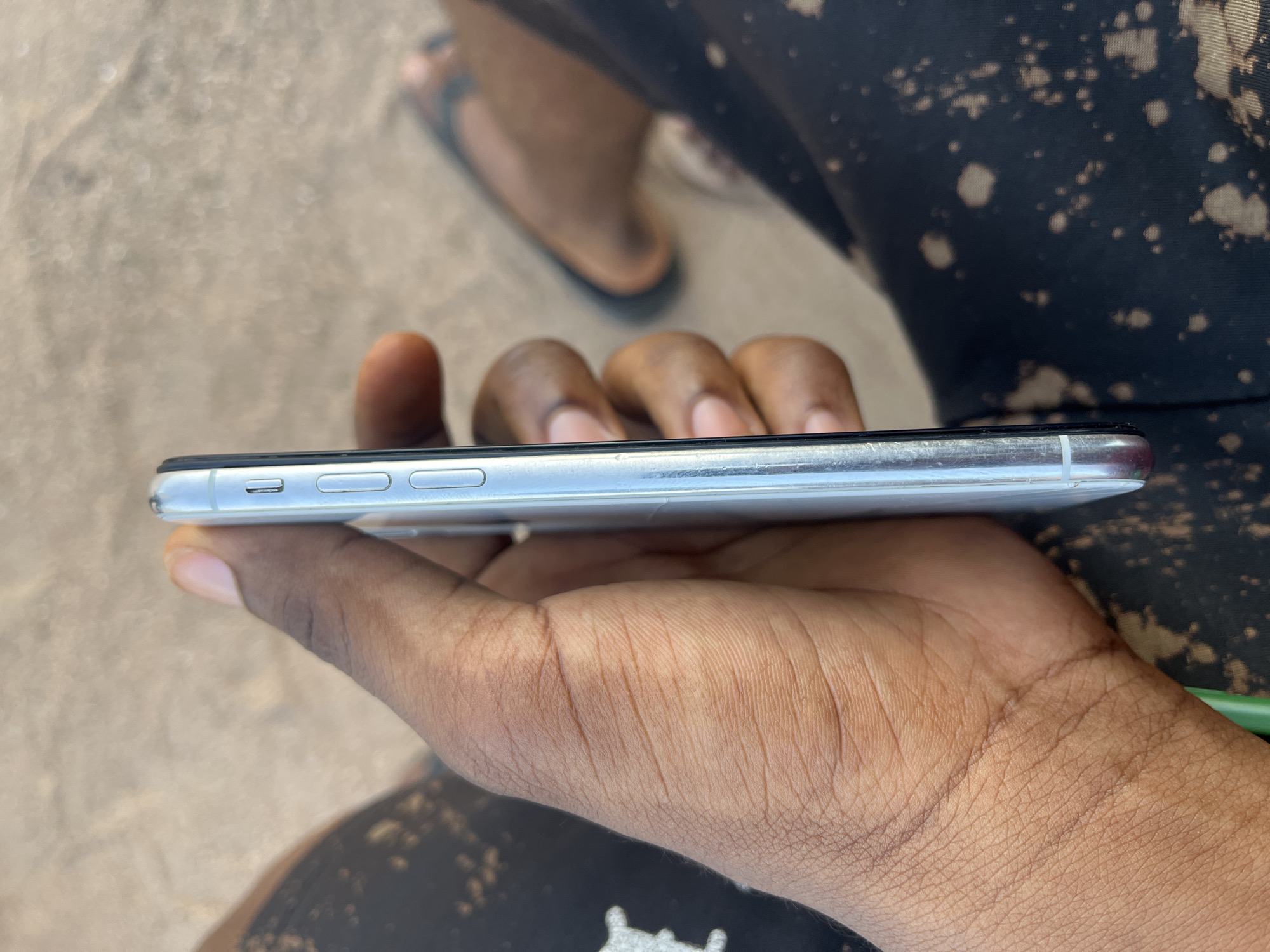 An African person's hand, with visible fingerprints, holds a silver smartphone vertically against their thigh. The phone's side buttons are visible. The person is wearing black shorts with a bleached pattern. The background is a sandy, light brown surface, possibly dirt or sand, with some blurred figures in the distance, one wearing sandals. The image appears to be taken outdoors in Jalingo, Nigeria.
