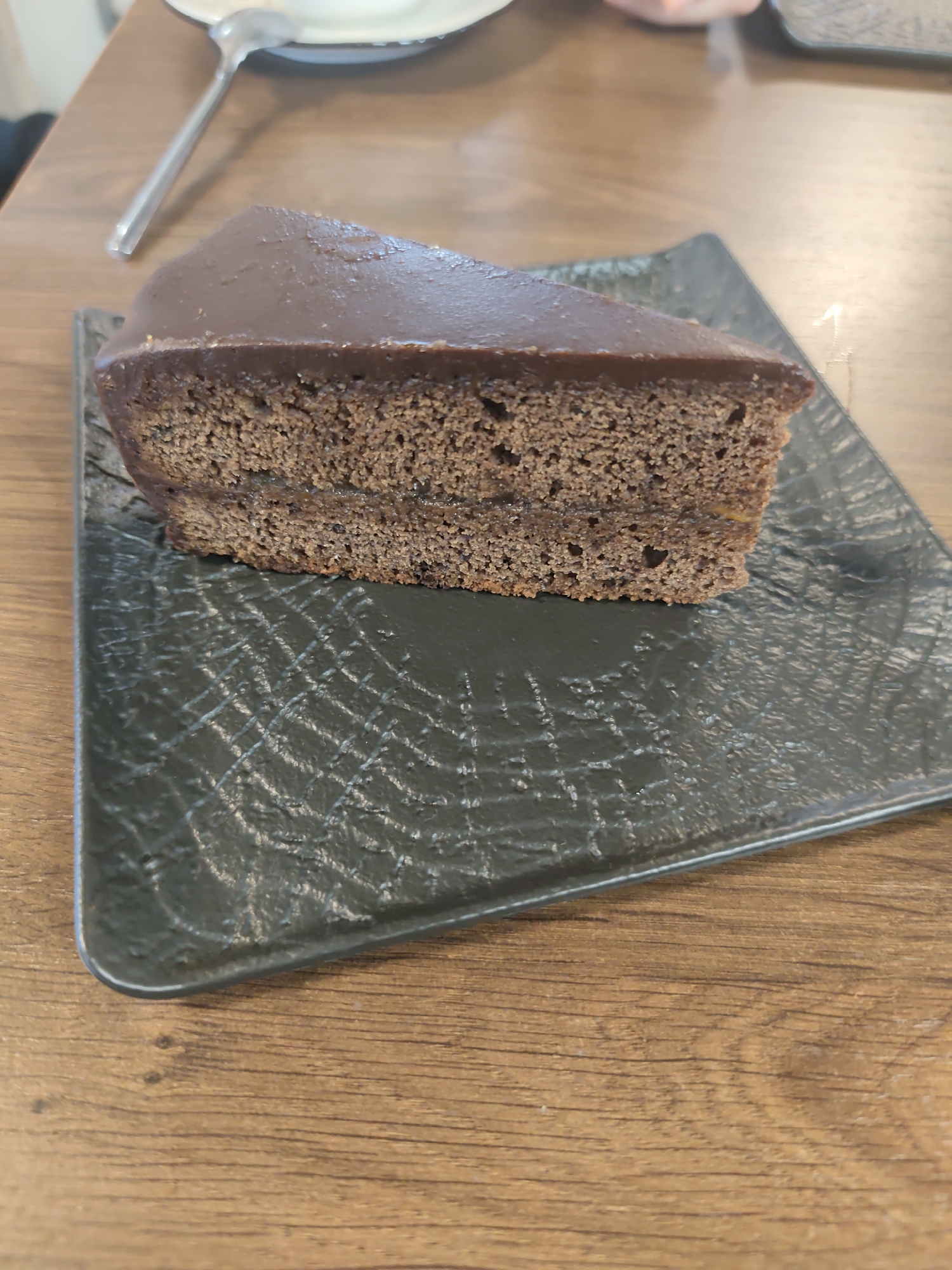 The image presents a close-up of a single slice of chocolate cake resting on a dark, square plate. The cake itself is dark brown, featuring a dense, porous crumb, suggesting a moist texture. It appears to be a layered cake, with a thin, lighter-colored filling, possibly apricot jam, visible between the two cake layers. The entire slice is coated with a smooth, glossy layer of dark chocolate ganache or icing.
The cake is positioned diagonally on a black or very dark gray plate with a textured surface. The plate has an intricate, somewhat irregular pattern of etched or embossed lines, resembling a web or a cracked surface, which reflects light subtly.
The plate and cake are situated on a wooden table with a light to medium brown tone and a visible wood grain pattern. In the blurred background, parts of a silver spoon and the edge of a white ceramic dish are discernible, implying a dining or cafe setting. The lighting is soft and even, typical of an indoor environment, with no harsh shadows or indications of a specific time of day. There are no individuals or visible text in the image. The scene captures a dessert moment, with the location context provided as Chaikovsky, Russia.