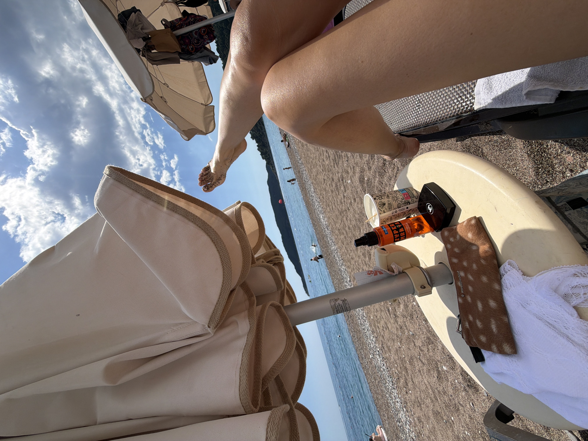 A first-person perspective shows a beach scene with a view of the Adriatic Sea in Boreti, Montenegro. The observer sits under a beige beach umbrella. Bare legs, glistening with oil, extend from the upper right. On a small, round table attached to the beach chair are a cup, an orange sunscreen bottle, a black cosmetic compact (likely Chanel), and a brown and white speckled glasses case next to a white cloth. The beach, strewn with pebbles, slopes down to the sea. Several people are scattered along the shoreline and in the water. A sliver of forested hill is visible on the left horizon. The sky is blue with scattered clouds.