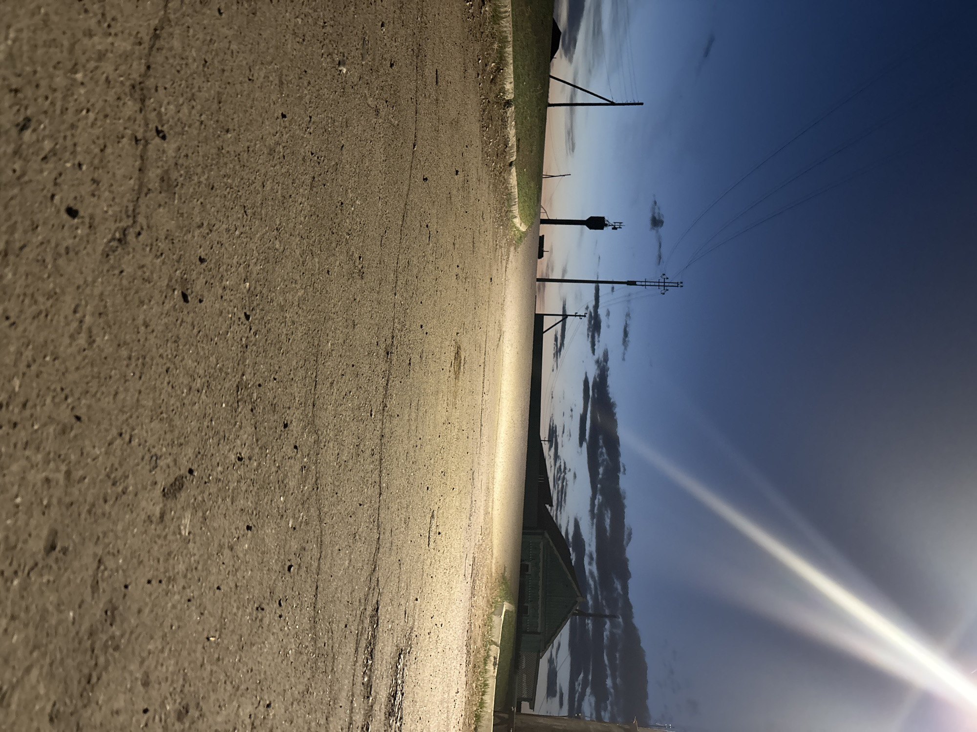 The image depicts a rural road, likely in Zagatuy, Russia, given the presence of utility poles and a water tower. The road surface is paved and rough, showing signs of wear. Adjacent to the road is a narrow strip of vegetation. Utility poles with power lines stretch into the distance, against a backdrop of a dusk or dawn sky with clouds. A building is also visible. The sky is illuminated by a bright light source, obscuring some details.