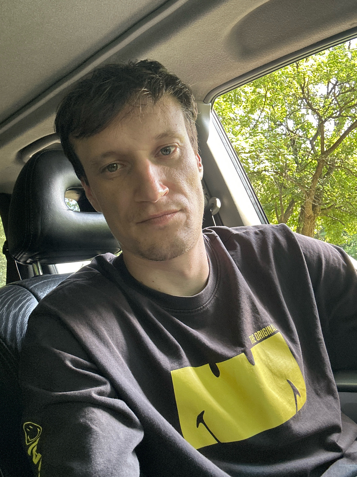 A man is sitting in the driver's seat of a car, looking at the camera. The man has short, dark hair and light-colored eyes. He is wearing a dark gray t-shirt with a large yellow smiley face logo on the front. The interior of the car is visible, including the headrest of the passenger seat and a portion of the car's ceiling. Outside the car window, green trees are visible, suggesting an outdoor location. The image does not contain any information that would help to identify the specific location in the city of Shatava, Ukraine.