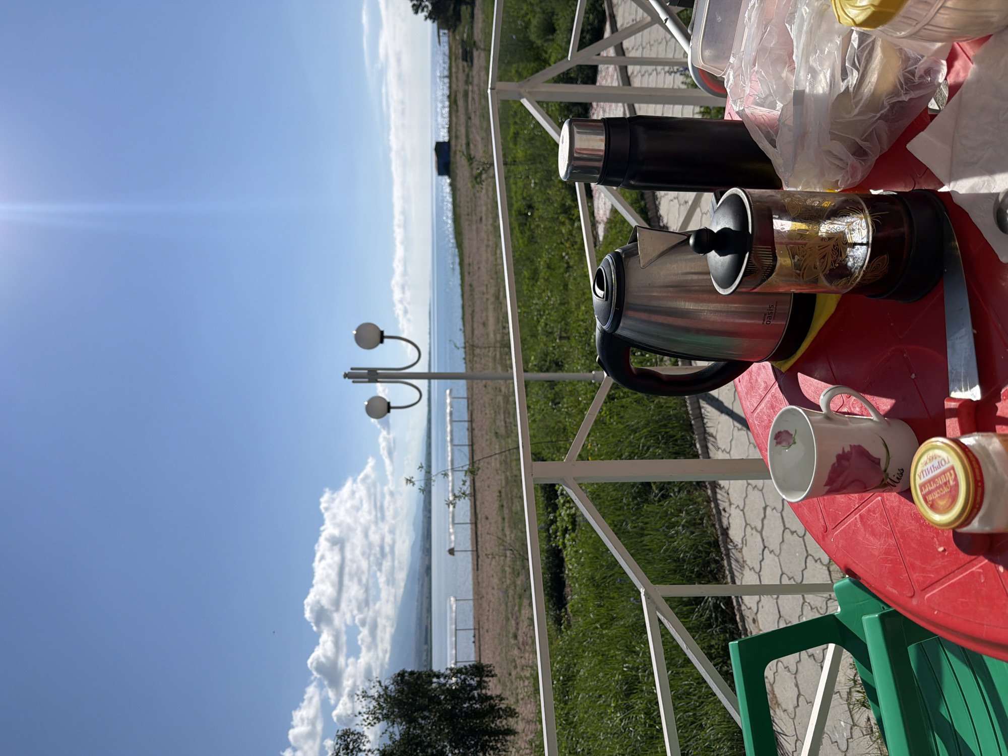 A high-angle, close-up shot shows a red plastic table set up on a balcony, overlooking a tranquil body of water. The table is adorned with breakfast items, including a silver electric kettle, a black thermos, a French press filled with dark liquid, a white mug with a pink rose design, a jar with a red and yellow label, and a clear plastic bag containing food. A knife rests on the table, suggesting a meal is in progress.
The balcony is bordered by a white metal railing, beyond which stretches a sandy beach and a grassy area. In the distance, the calm water of a lake or sea meets a hazy shoreline, with mountains forming a distant backdrop under a bright blue sky dotted with scattered white clouds. A lamppost with two spherical lights stands near the railing, adding a touch of rustic charm to the scene. The overall impression is one of a serene breakfast enjoyed with a picturesque view.