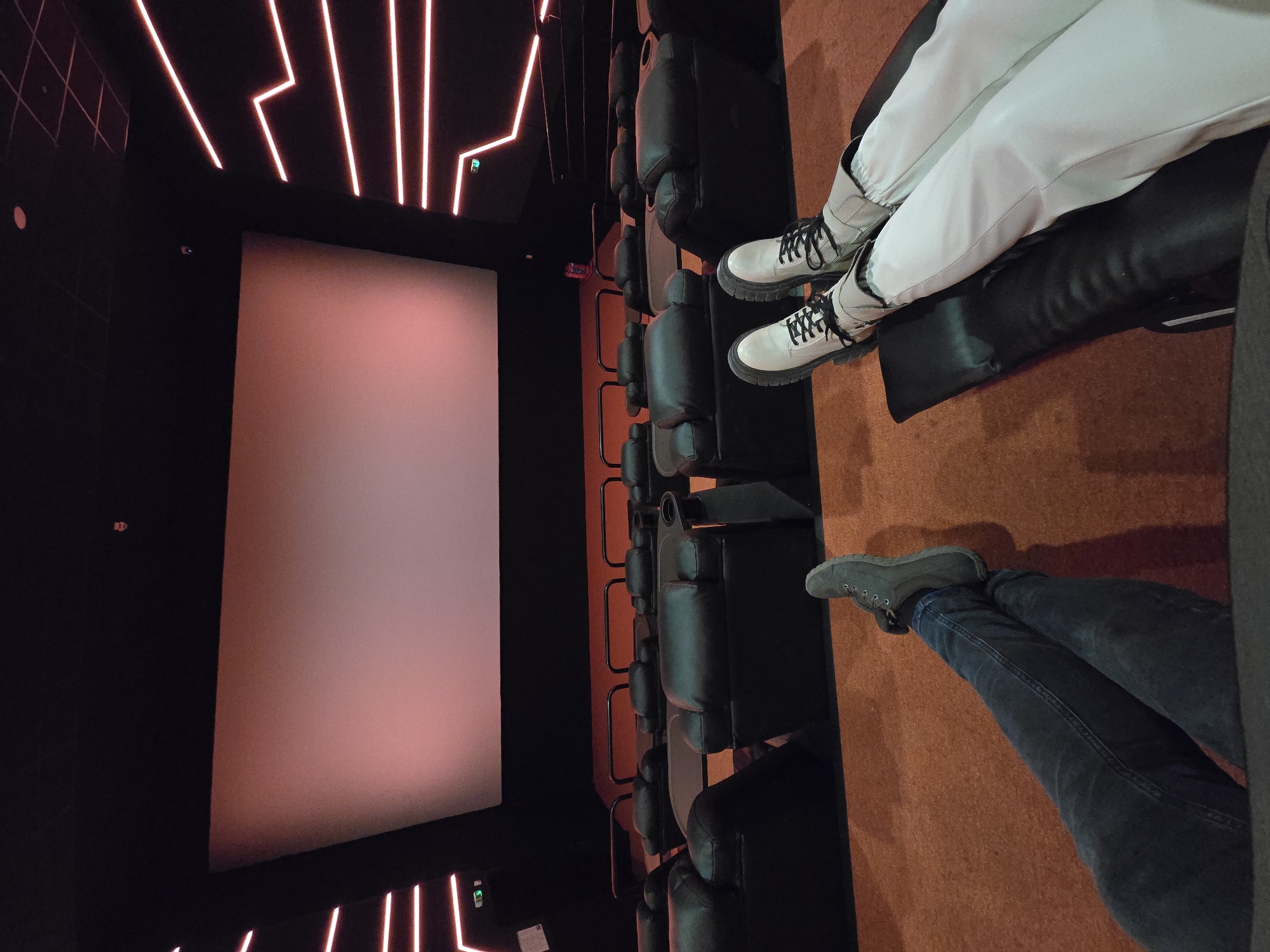 The image depicts the interior of a cinema hall in Varna, Bulgaria, observed from a seated, low-angle perspective. In the immediate foreground, the extended lower legs of two individuals are visible. On the left, a person wears dark blue jeans and grey, possibly suede, boots with black laces, their legs crossed at the ankles. On the right, a person wears light-colored trousers and light beige, thick-soled boots with black laces, resting their legs on a black reclining footrest.
Beyond the foreground subjects, multiple rows of unoccupied black, padded reclining cinema seats are visible, each featuring a cup holder on the armrest. The floor is covered with a reddish-brown carpet. The central background is dominated by a large, rectangular cinema screen, emitting a soft, uniform reddish-pink illumination. Dark walls frame the screen, accented by horizontal red LED or neon light strips arranged in angular, geometric patterns. A green illuminated 'EXIT' sign is affixed to the lower left wall. A small red object, potentially a fire extinguisher, is visible on the floor behind a row of seats towards the right side of the screen. The dark ceiling features a visible grid pattern, and a small, dark security camera is mounted centrally near the top of the frame.