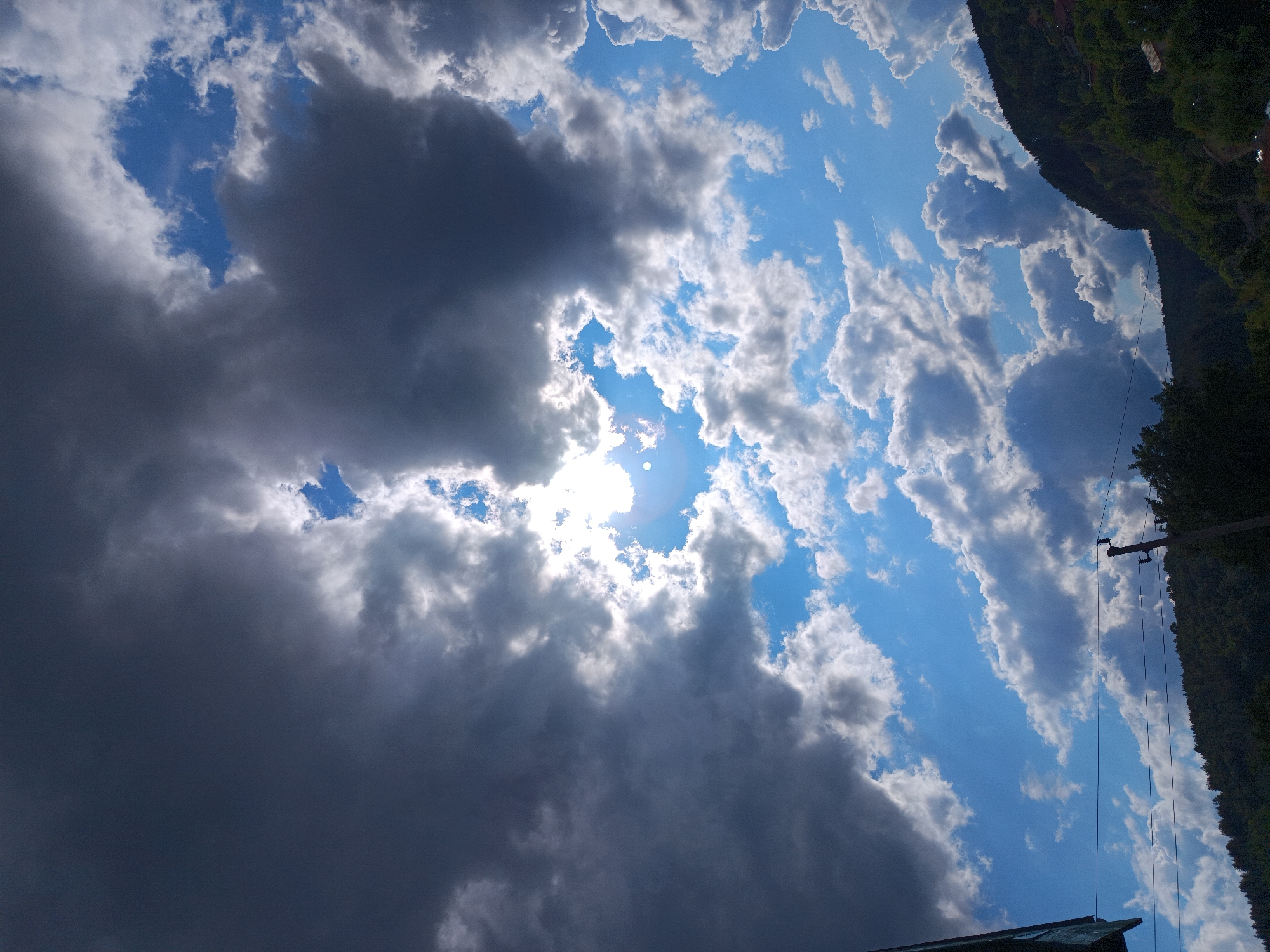This media file captures a dramatic sky scene with a mix of dark, heavy clouds and bright, sunlit patches. The sun is visible through a break in the clouds, creating a radiant glow and lens flare. The clouds are voluminous and textured, suggesting active weather conditions, possibly an approaching storm or the clearing after one. The sky is a vibrant blue in the areas not obscured by clouds.
To the right, the image shows the dark green slope of a mountain or hill covered in dense trees. The terrain appears steep, indicating a mountainous or hilly region. A power line with insulators and a utility pole runs vertically along the edge of the tree line, suggesting some human infrastructure in the otherwise natural landscape. At the very bottom, a dark-colored roofline of a building is visible, hinting at a settlement or dwelling nearby. The perspective of the shot is from below, looking upwards, which emphasizes the scale and grandeur of the sky.
The lighting suggests it is daytime, likely mid-afternoon given the intensity of the sun and the shadows cast by the clouds. The overall mood is one of dynamic natural beauty, with a touch of dramatic intensity due to the contrasting light and dark clouds. The location context of Kralev Dol, Bulgaria, aligns with the mountainous terrain and forested slopes. There is no visible text in the image.