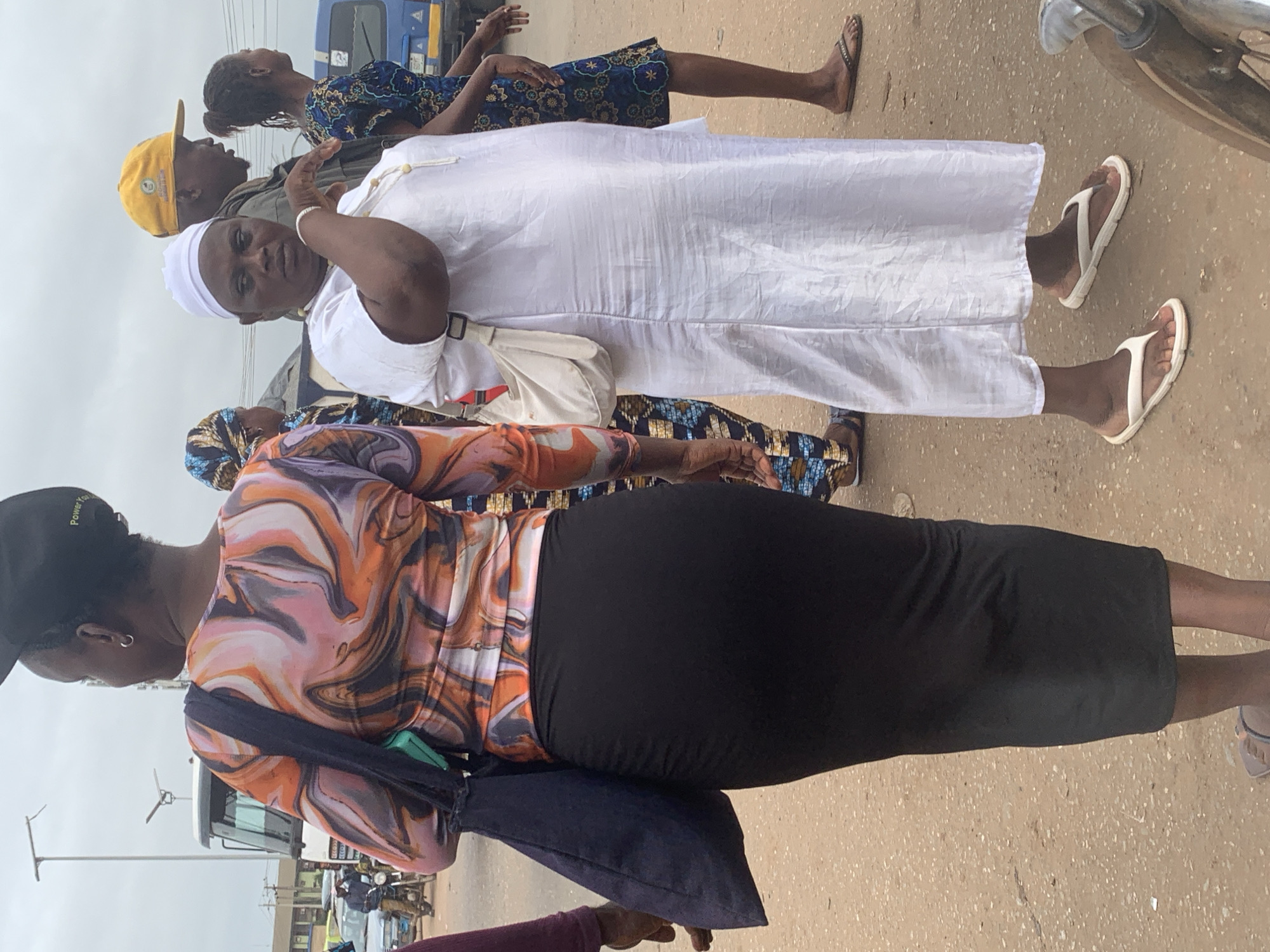 In the city of Shagamu, Nigeria, three women and one man are visible outdoors. The foreground is occupied by a woman wearing a black, form-fitting skirt and a long-sleeved shirt with a swirling orange, pink, and black pattern. She carries a dark blue tote bag over her left shoulder. To her right, a woman in a white robe and head covering stands with her right hand raised, touching her chin. She wears a light-colored crossbody bag. Behind her, a man in a yellow baseball cap is seen from the side, and further to the right, another woman in a blue and yellow patterned dress walks away from the camera. The ground is covered in dirt and gravel, with a hint of a paved surface. In the background, a white bus and other vehicles are visible, suggesting a street or transportation hub. The sky is overcast.