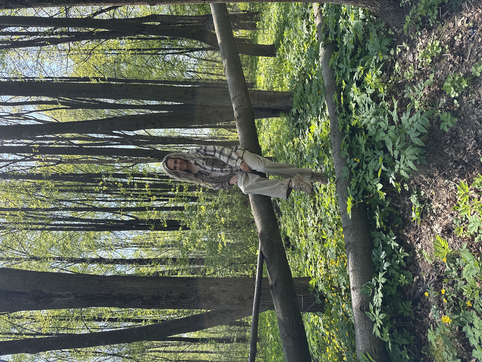 The image shows a woman sitting on a tree trunk in a forest or wooded area. She has blonde hair and is wearing a plaid shirt, light-colored pants, and shoes. The trees are tall and thin, and there is green foliage on the ground. The scene appears to be outdoors in natural light. The location mentioned, Rusanovo, Russia, cannot be independently verified from the image.