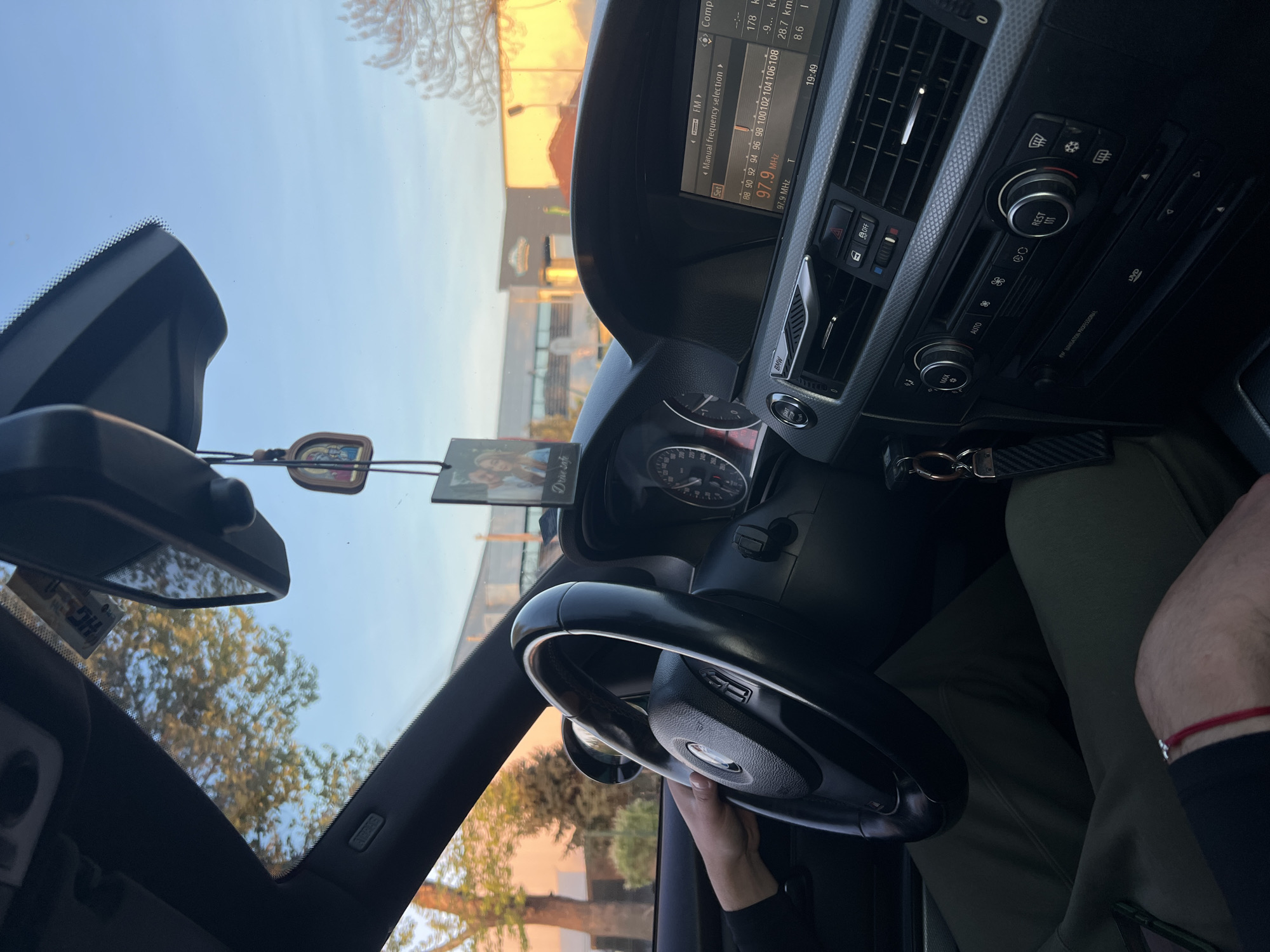 The image captures the interior of a car, likely a BMW given the visible logos and dashboard design, from the perspective of the passenger seat looking towards the driver. A person's right hand is visible holding the steering wheel. The driver is wearing dark green pants, and a red string bracelet is visible on their right wrist.
The dashboard features a prominent infotainment screen displaying radio information, specifically tuned to "97.9 MHz FM," and the time "19:49." Other readouts on the screen include "Comp" (presumably "Computer"), "178 k," "28.7 km," and "8.6." Below the screen are air vents and climate control buttons, including "MAX," "AUTO," "REST," and buttons for fan speed and air direction. A "START/STOP" engine button is also visible. The "DVD" slot and text "NAVIGATION PROFESSIONAL" further identify the car's features.
Through the windshield, a clear blue sky and warm, soft sunlight are visible, suggesting it is late afternoon or early evening. Trees with yellow-green foliage and some distant buildings or structures are seen outside. A small charm or air freshener hanging from the rearview mirror features the text "Drive safe" and an image of people.
The scene depicts someone driving or about to drive a car in Plovdiv, Bulgaria, during the evening hours.