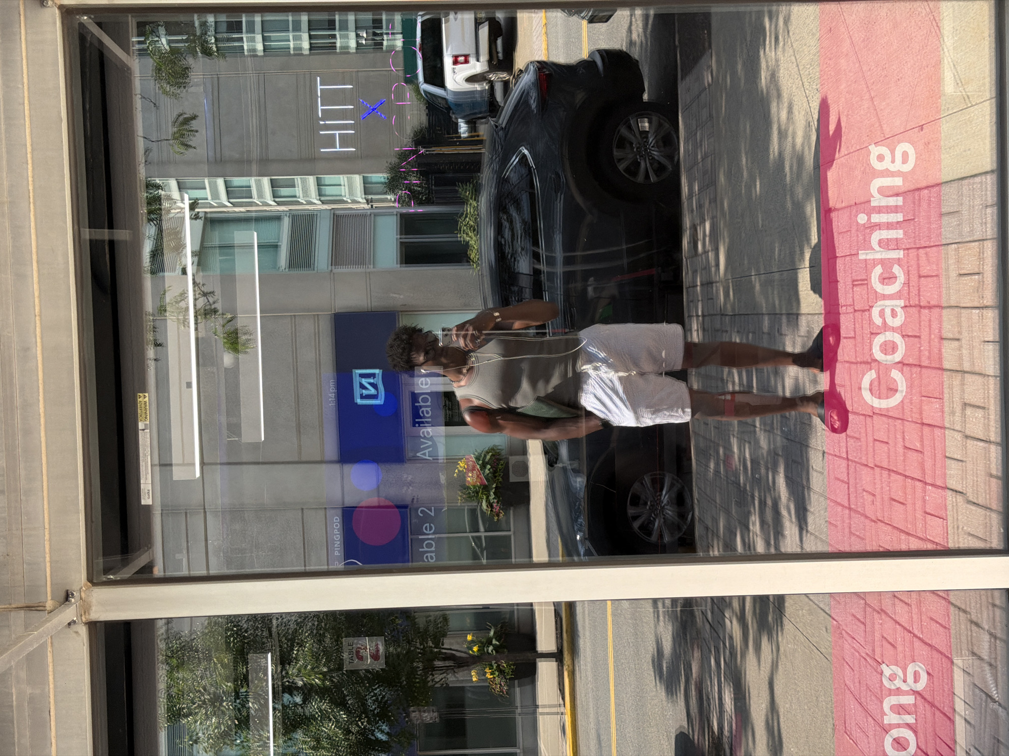 In Jersey City, New Jersey, United States of America, a young Black male is captured in a mirror selfie taken at a bus stop. He stands in front of a reflective glass panel, wearing a grey tank top, white shorts, and dark slides. His hair is styled in short locs, and he sports a chain necklace and earrings. He is holding a smartphone in his right hand, angled to capture his reflection.
The bus stop's glass reflects the urban environment. Across the street, buildings with modern architecture are visible, including one with prominent neon signage that reads "HITT X". The street is lined with trees and potted plants, and a dark-colored SUV is parked on the side. In the foreground, the glass displays text in pink, with "Coaching" partially visible, suggesting the location is a business or service center. The ground under the bus stop is paved with interlocking bricks. The scene is bathed in bright sunlight, casting shadows on the sidewalk.