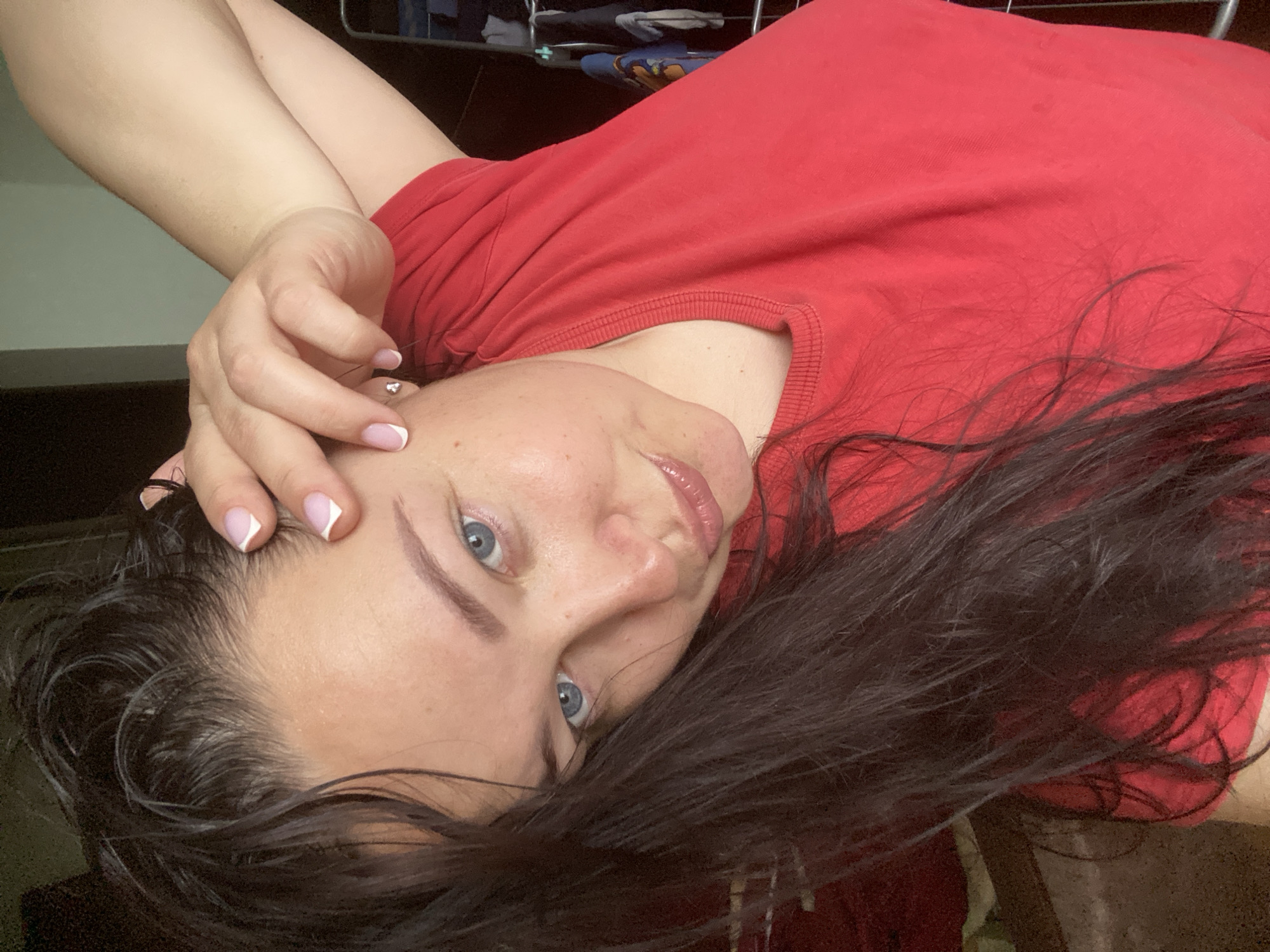A woman with long, dark hair and blue eyes is reclining, wearing a red t-shirt. Her left hand is near her head, displaying a French manicure. The background is dark and indistinct, but includes a wire shelf. The image does not provide sufficient information to determine a location in Sochi, Russia.