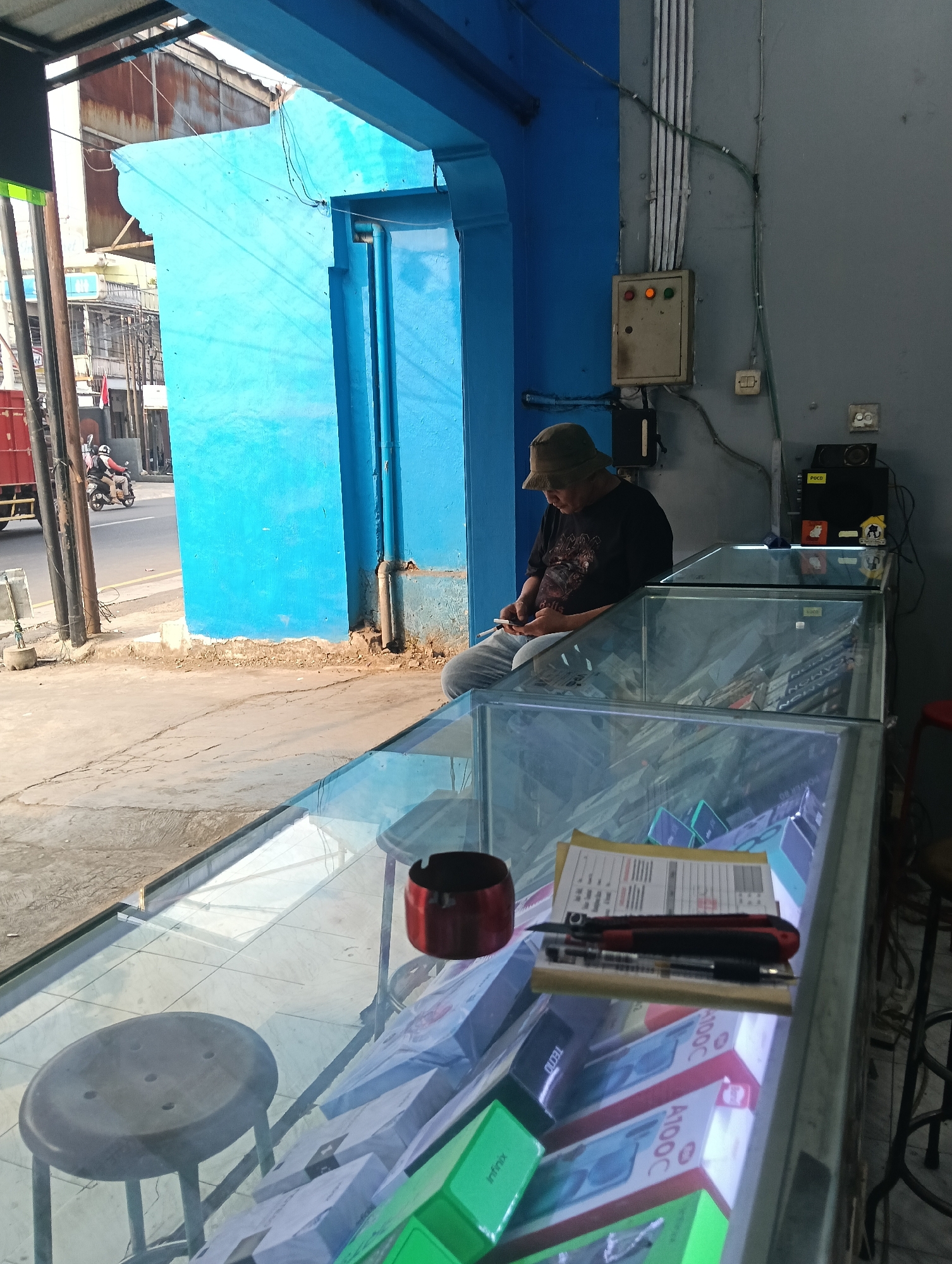 A middle-aged man wearing a bucket hat and a dark t-shirt sits on a stool, looking down at his mobile phone. He is inside a shop with glass display cases filled with boxed items, possibly electronics, such as phones. A red ashtray and a utility knife with a pen are placed on top of a document on one of the display cases. The shop has a bright blue wall and entrance area. Outside, a street is visible with a red truck and a person riding a motorcycle. The scene appears to be in Desa Cipakat, Indonesia, judging by the context provided.