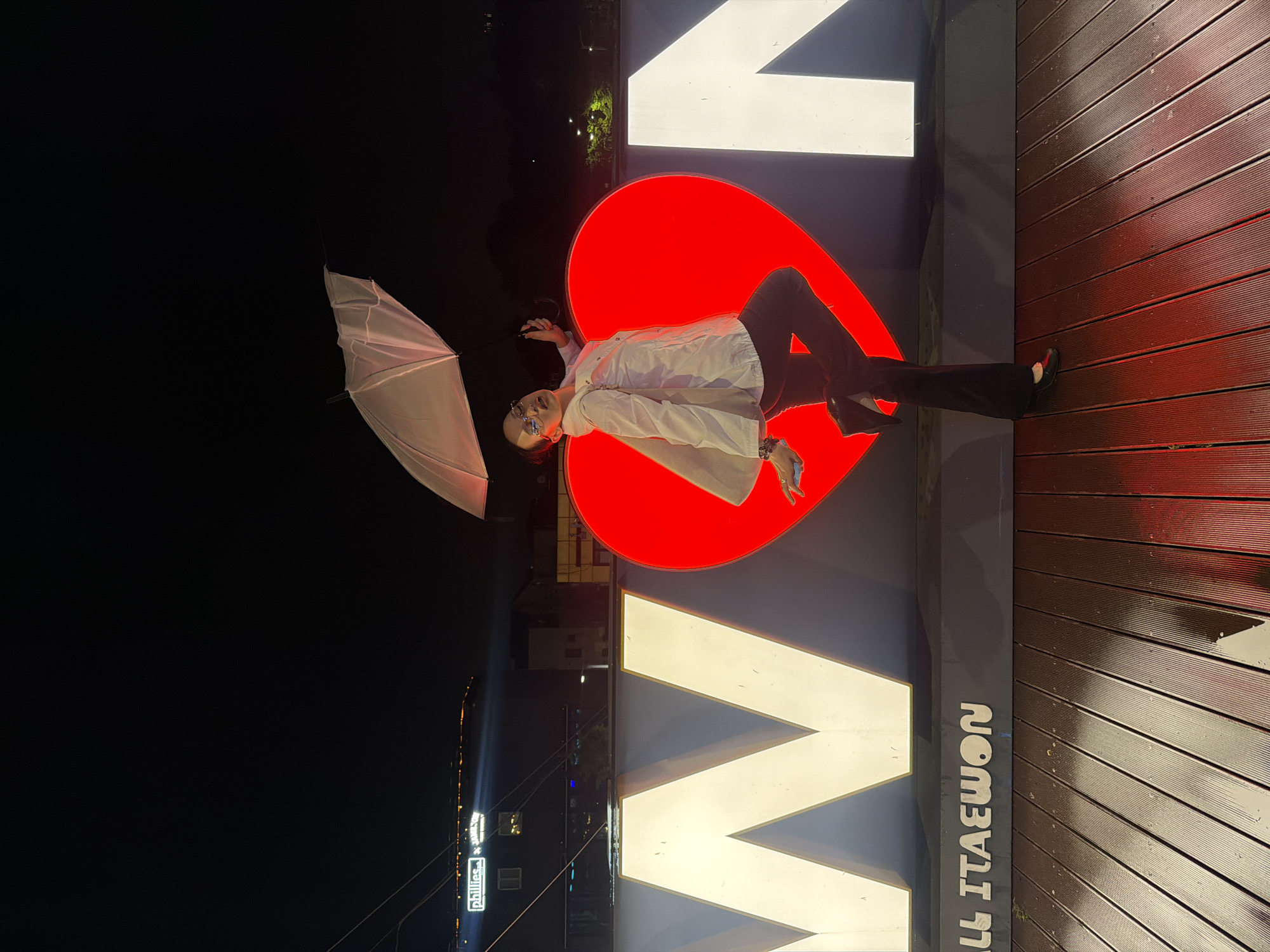A person lies angled, wearing a light-colored shirt, dark pants, and shoes, positioned on a large sign featuring a red heart and the word "ITAΕWON" in white letters. The location is Itaewon, a district in Seoul, South Korea. The person holds an umbrella, and the scene is set at night, with a wooden surface visible.