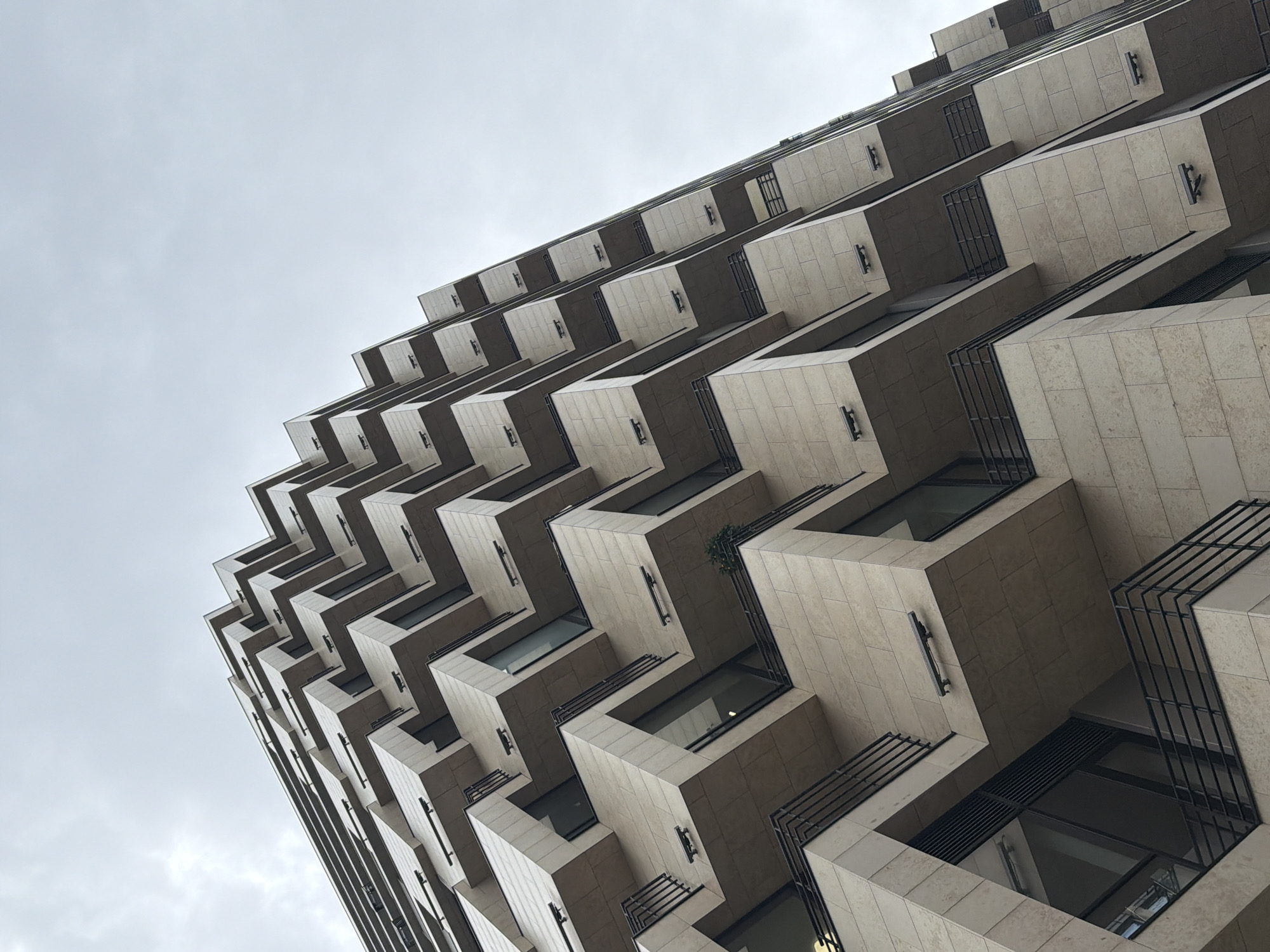A low-angle shot looks up at a multi-story building with an architectural design of stacked, cantilevered balconies. The building is constructed of light beige or cream-colored stone tiles and each balcony has a dark, metal railing. The building's shape gives it a distinctive zig-zag appearance. The sky is overcast and grey. The image is taken in Moscow, Russia.