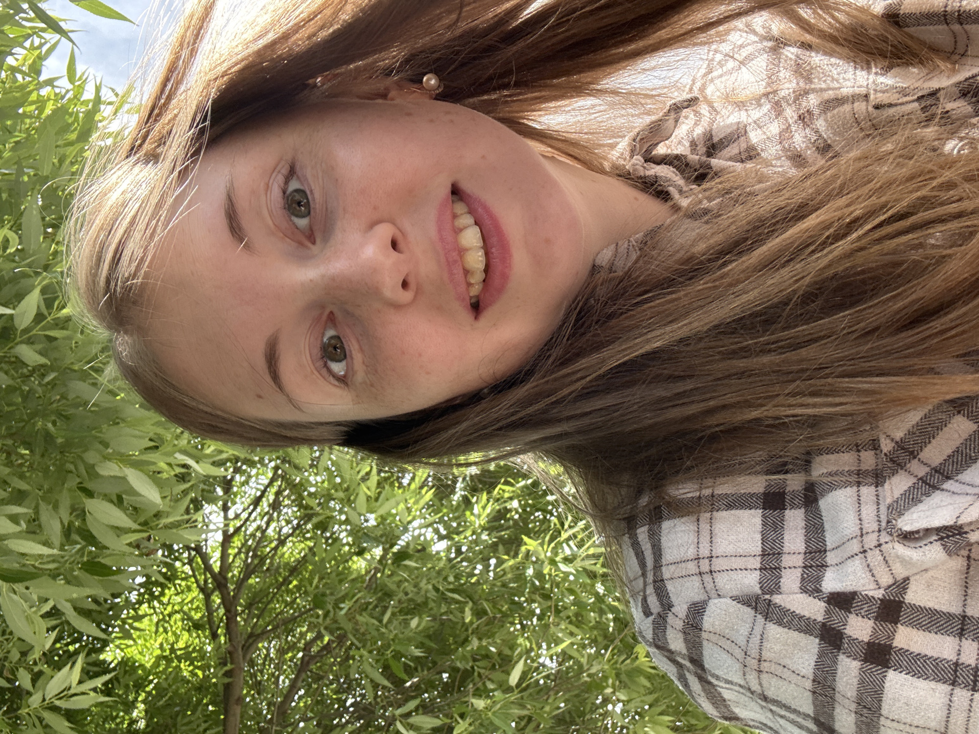 A selfie of a young woman with long brown hair and fair skin with freckles is shown. She is wearing a plaid shirt. She is looking at the camera and smiling. The background of the image is a tree with green leaves. The lighting suggests it is daytime.