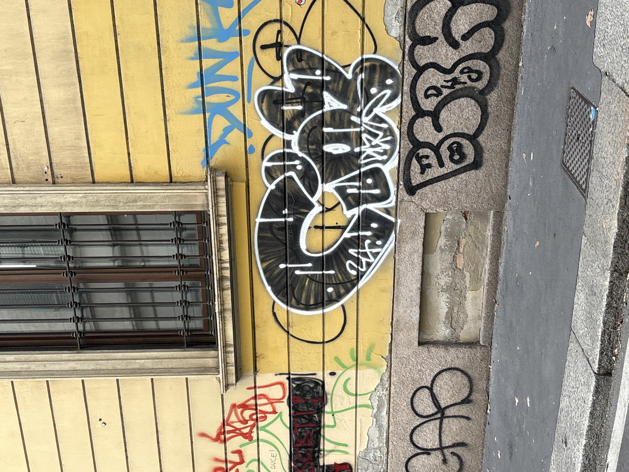 The image shows a section of a building in Milan, Italy, featuring a mustard-yellow, vertical-plank wall marred by various graffiti tags. A window with metal bars is present on the left side of the wall. Next to the window, large, black graffiti bubble letters with white outlines form an illegible word. Adjacent to this, other black graffiti tags are visible on a concrete section of the building. Below the graffiti is more graffiti in red and green, and an additional window/ventilation covering. To the right is a sidewalk and curb.