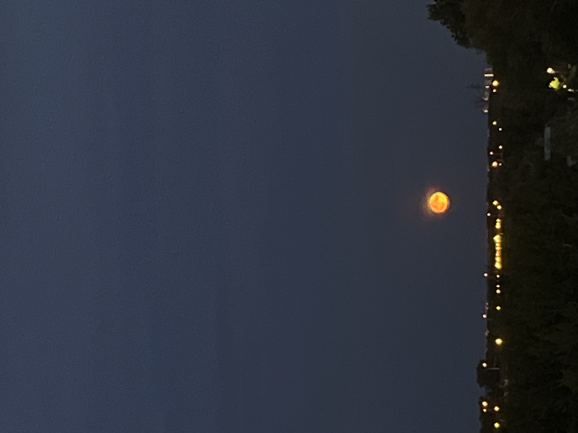 The image shows a dark blue sky with a large, orange moon visible in the upper center. Below the moon, a silhouette of trees and a city skyline with lights can be seen. The lights appear to be streetlights and building lights. The overall scene depicts a nighttime view of a city, possibly Samara, Russia, with a prominent moon.