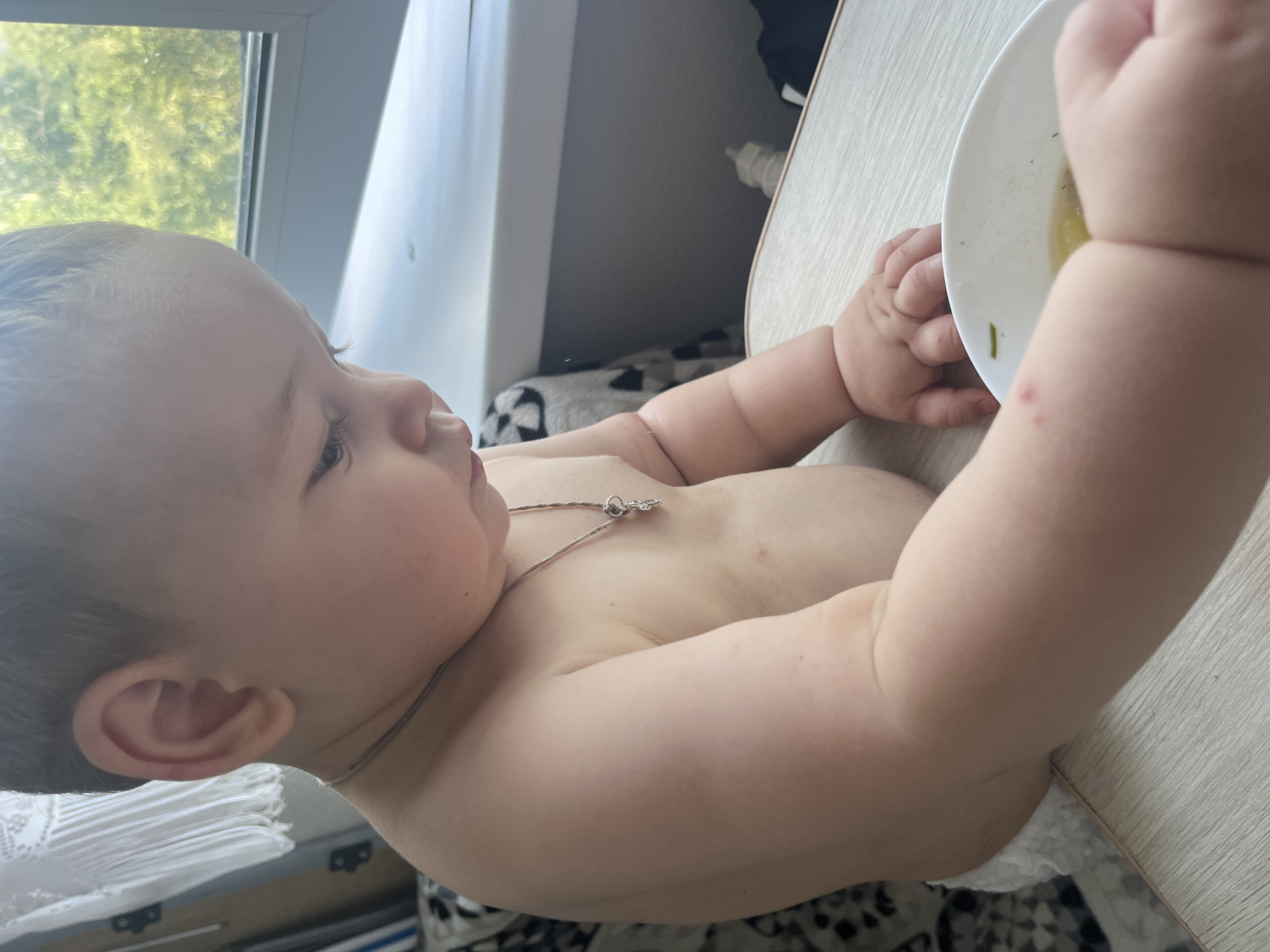 A close-up, eye-level, medium shot captures a baby from the chest up, seated at a light-colored table. The baby is facing right, looking down at a white bowl of food. The baby's skin appears fair, with a few red marks visible on the left arm. The baby is wearing a thin silver necklace with a small cross pendant. The background shows a window with green foliage visible outside, suggesting an outdoor environment. The room appears to be a domestic setting, possibly a dining area. No specific landmarks identifying Chelyabinsk, Russia are discernible in the image.
