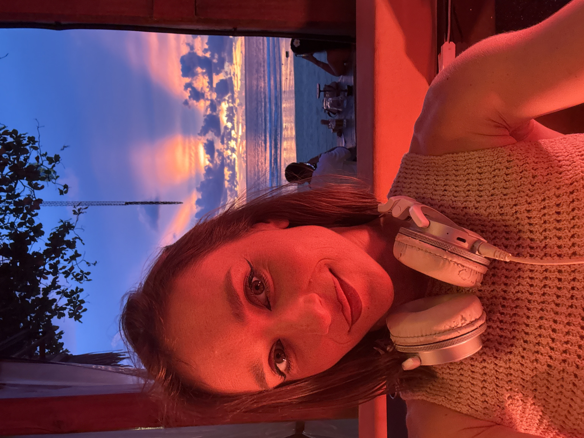 A woman is captured in a close-up, sideways shot, her face illuminated by warm, red lighting. She has brown hair and is wearing a light-colored, textured sweater. A pair of white headphones rests on her shoulder, one earcup partially visible. Her expression is a slight smile, with her eyes looking towards the camera.
Behind her, a window or opening reveals a stunning sunset over the ocean. The sky is a vibrant mix of deep blue, purple, and fiery orange and pink hues, with scattered clouds. The ocean reflects the colors of the sky, creating a serene backdrop. In the distance, silhouetted figures can be seen near the water, and a tall, dark structure, possibly a tower or crane, stands against the twilight sky on the left side of the frame. A hint of leafy branches from a tree is also visible on the far left.
The overall mood of the image is peaceful and relaxed, with a sense of enjoying a beautiful natural spectacle. The red lighting on the woman suggests an artificial light source within the immediate foreground, possibly from the location she is in. No visible text is present in the image. The time of day is clearly sunset.