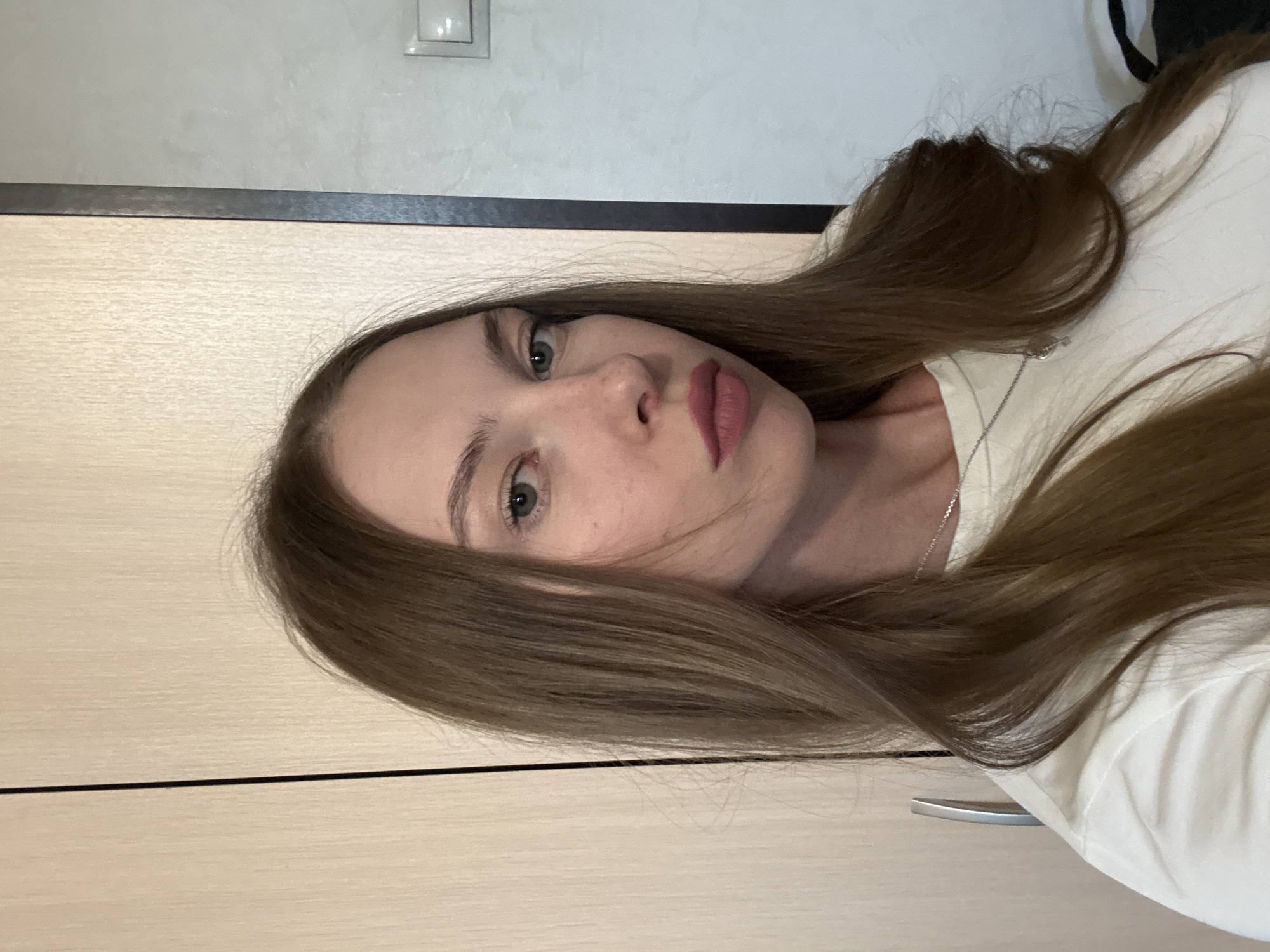 This is a close-up, vertically oriented photograph of a young woman. She has long, light brown hair that is falling across her face and shoulders. Her eyes are a light green, and she has fair skin with some freckles visible. She is wearing a white t-shirt and a delicate silver necklace. The background consists of a light-colored, textured wall and a wooden surface, possibly a cabinet or door, with a metal handle. A thin black line runs horizontally across the image, separating the wall from the wooden surface. There is a light switch on the wall. The overall lighting suggests an indoor setting, possibly during the daytime. The woman's expression is neutral, and the image appears to be a selfie. No text is visible in the image.