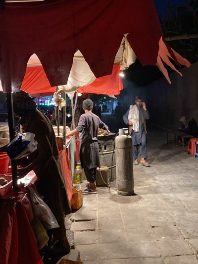 The image shows a nighttime scene in Kandahar, Afghanistan, at a food stall. The stall is covered by a red and white awning. Two people are working at the stall; one wearing a head covering is preparing food, while the other is cooking over a stove next to a propane tank. Another man is standing nearby, talking on a cell phone while wearing a shawl. In the background, several people are sitting on a bench along a wall. The ground is paved with stone tiles.