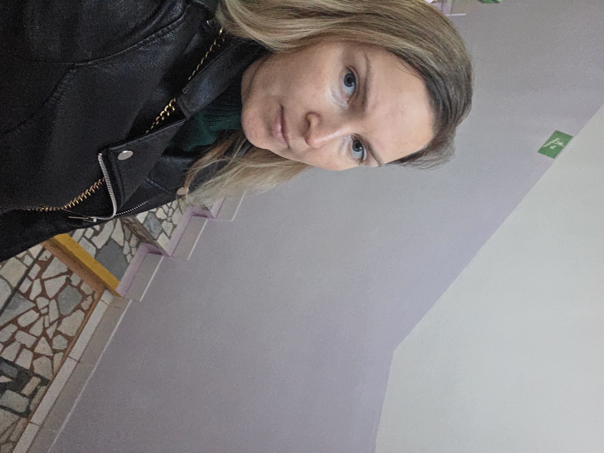 A woman with blonde hair and blue eyes is posing for a picture, wearing a black leather jacket over a green turtleneck sweater. She is standing inside a building, possibly in the city of Chaikovsky, Russia. The floor has a mosaic tile pattern, and there is a white wall with a lilac tint. There is a green sign on the wall in the upper right corner. The picture is taken at an angle.