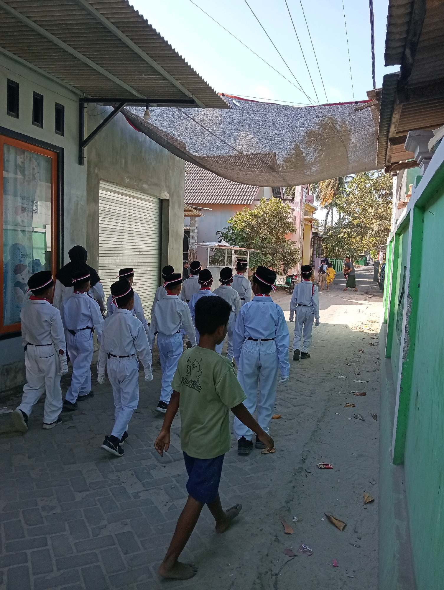 A group of young boys wearing white uniforms and black hats are walking down a narrow, sandy street in Pagerungan Kecil, Indonesia. The boys are part of a school or religious organization, and their attire suggests they are participating in a parade or ceremony. The street is lined with buildings, some of which are covered with corrugated metal roofs. In the background, there are palm trees and other vegetation, indicating a tropical climate. The sky is clear and blue, suggesting a sunny day. The scene appears to be a typical day in a small Indonesian village.