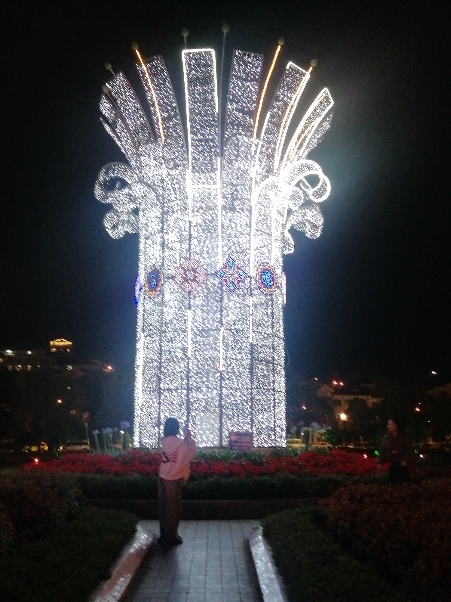 The image displays a vibrant nighttime scene centered around a large, elaborate light sculpture. The main subject is a towering, tree-like structure covered in countless small, bright white LED lights, illuminating the dark surroundings. This festive display features prominent vertical segments, topped with gracefully arching and fanning elements, all intricately lit. Embedded within the structure are several circular, colorful decorative motifs, appearing as mandalas or starbursts, featuring shades of blue, red, and yellow.
In the mid-ground, a person is visible from behind, observing the illuminated display. They are wearing a light-colored (possibly pink or beige) long-sleeved top and dark pants. Another figure is partially visible further to the right. The base of the light sculpture is encircled by neat flower beds filled with vibrant red and green foliage, separating the paved walkway in the foreground from the main attraction.
The background reveals other distant buildings, faintly lit with warm yellow lights, and streetlights, indicating an urban or town setting. The deep darkness of the sky confirms it is nighttime. The scene suggests a public space in Sa Pa Ward, Vietnam, where people are enjoying a festive or decorative light installation. A small, rectangular sign is visible at the base of the light structure, but its text is illegible. The overall mood is one of wonder and enjoyment.