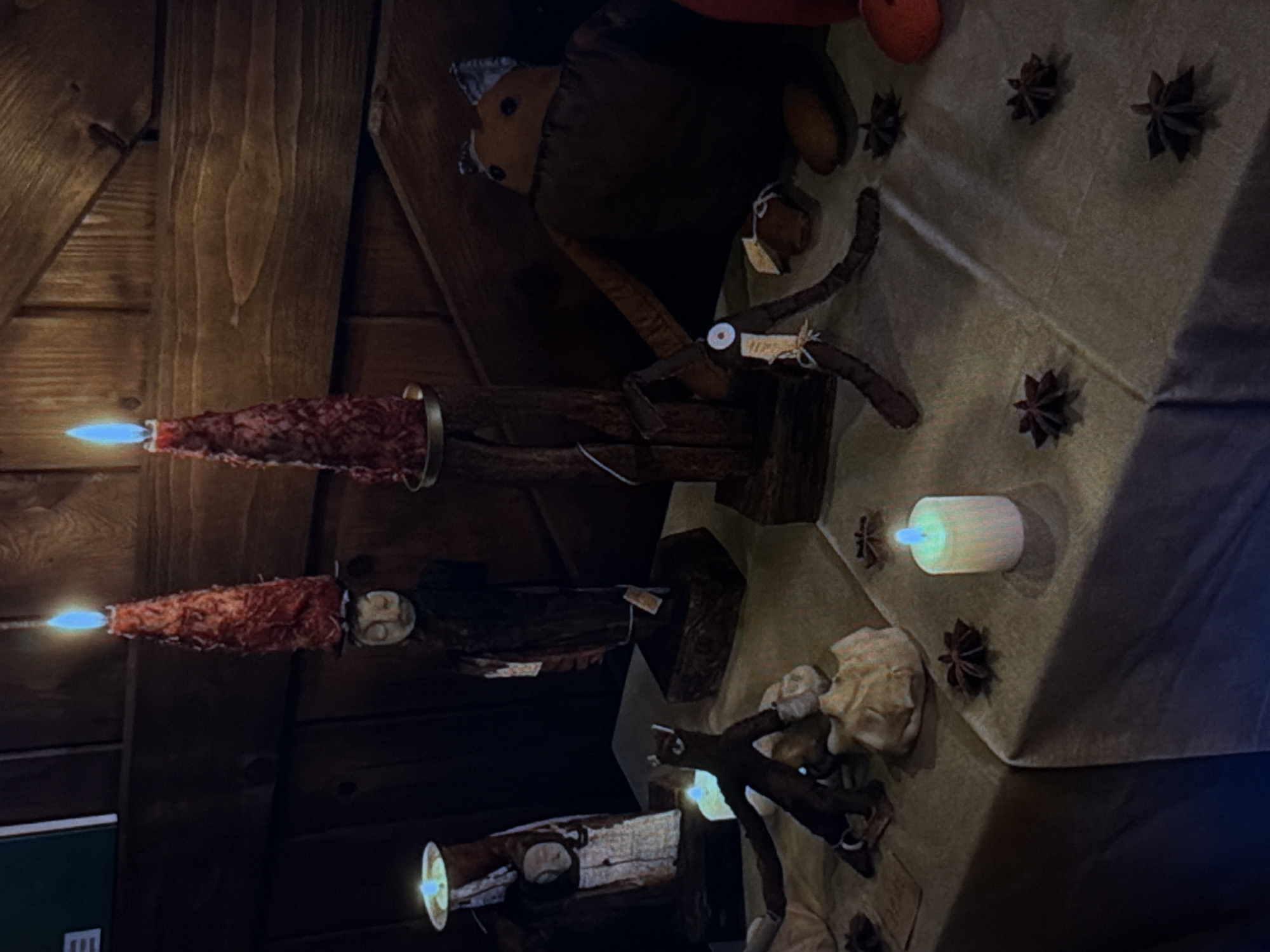 The scene depicts a dimly lit, rustic interior, possibly a cabin or shop, with wood paneled walls. Several lit candles are mounted on dark wooden blocks. The light emanates from the tops of the candles in a bright blue hue. The foreground features a table draped in a light-colored cloth, possibly velvet, adorned with star anise and what appear to be small dolls or figurines. A stuffed toy animal is also visible on the table. The location is not determinable from the image. There is no way to tell if this is Đà Nẵng, Vietnam from what is shown.
