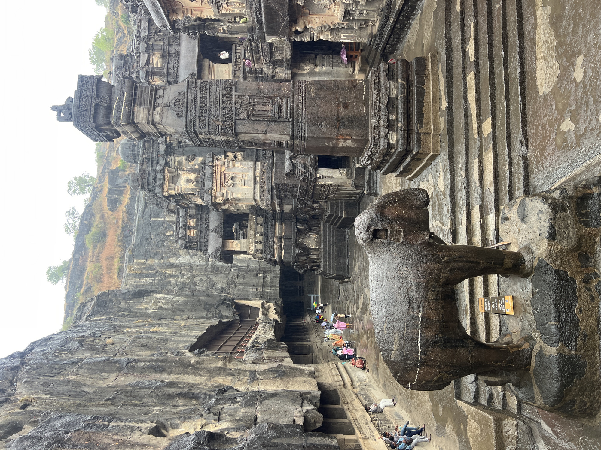 The image depicts the exterior of the Kailasa Temple at Ellora Caves, a UNESCO World Heritage Site in Maharashtra, India. The temple is carved from a single rock and features intricate sculptures and architectural elements. In the foreground, a large stone elephant statue stands on a platform. Several tourists, some with umbrellas, are visible exploring the complex, which appears to be wet, suggesting it is raining. The surrounding rock-cut architecture includes towering pillars and detailed carvings. The sky is overcast.