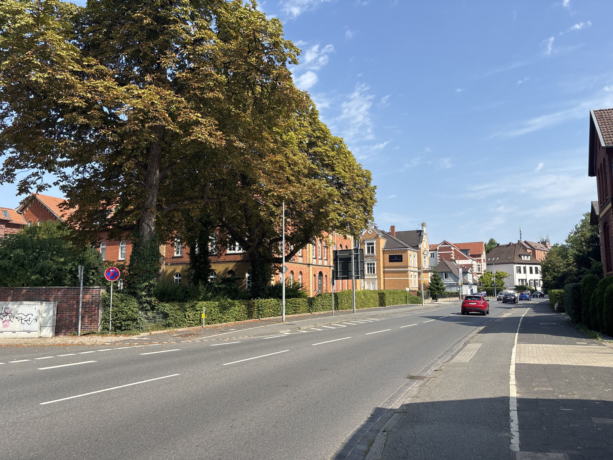 The image captures a street scene in Celle, Germany, on a clear, sunny day with a few scattered clouds. A wide asphalt road dominates the foreground, marked with white lane dividers. To the left, a large, mature tree with golden-brown leaves shades a brick wall adorned with graffiti and a white utility box. Beyond the wall and tree, a multi-story brick building with arched windows is visible, surrounded by dense green foliage. A red and white circular traffic sign with a diagonal red bar and a symbol of a car is posted on a metal pole.
The right side of the road features a sidewalk with a contrasting light-colored paved section and a dark asphalt section. A row of residential buildings with red-tiled roofs and a variety of architectural styles lines the street, interspersed with green hedges. Several cars are present on the road, including a prominent red SUV in the middle distance, traveling away from the viewer, and other vehicles further down the street. The overall impression is of a residential or mixed-use area in a German town.