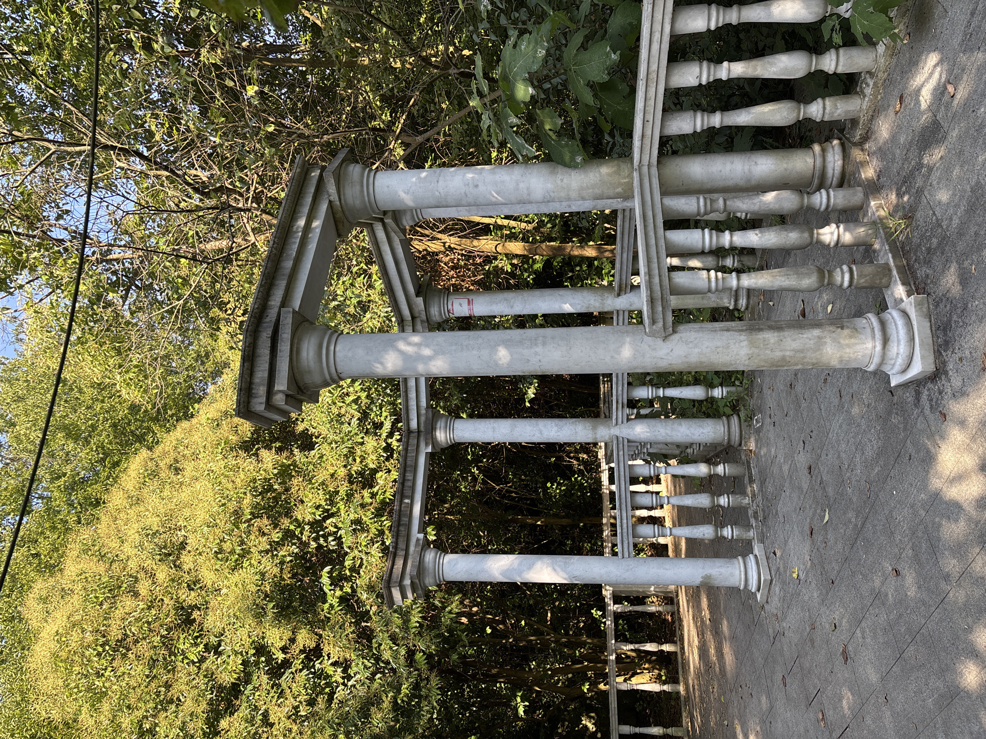 This is a medium-angle, eye-level shot of a white marble gazebo located in a park in Sochi, Russia. The gazebo features classical columns and railings and is surrounded by lush green trees. The ground in front of the gazebo is paved with gray tiles and scattered with fallen leaves. The lighting suggests it is daytime, with sunlight casting shadows on the ground. No people are visible in the image. The environment is serene and natural, with a sense of historical architecture integrated into a natural landscape.