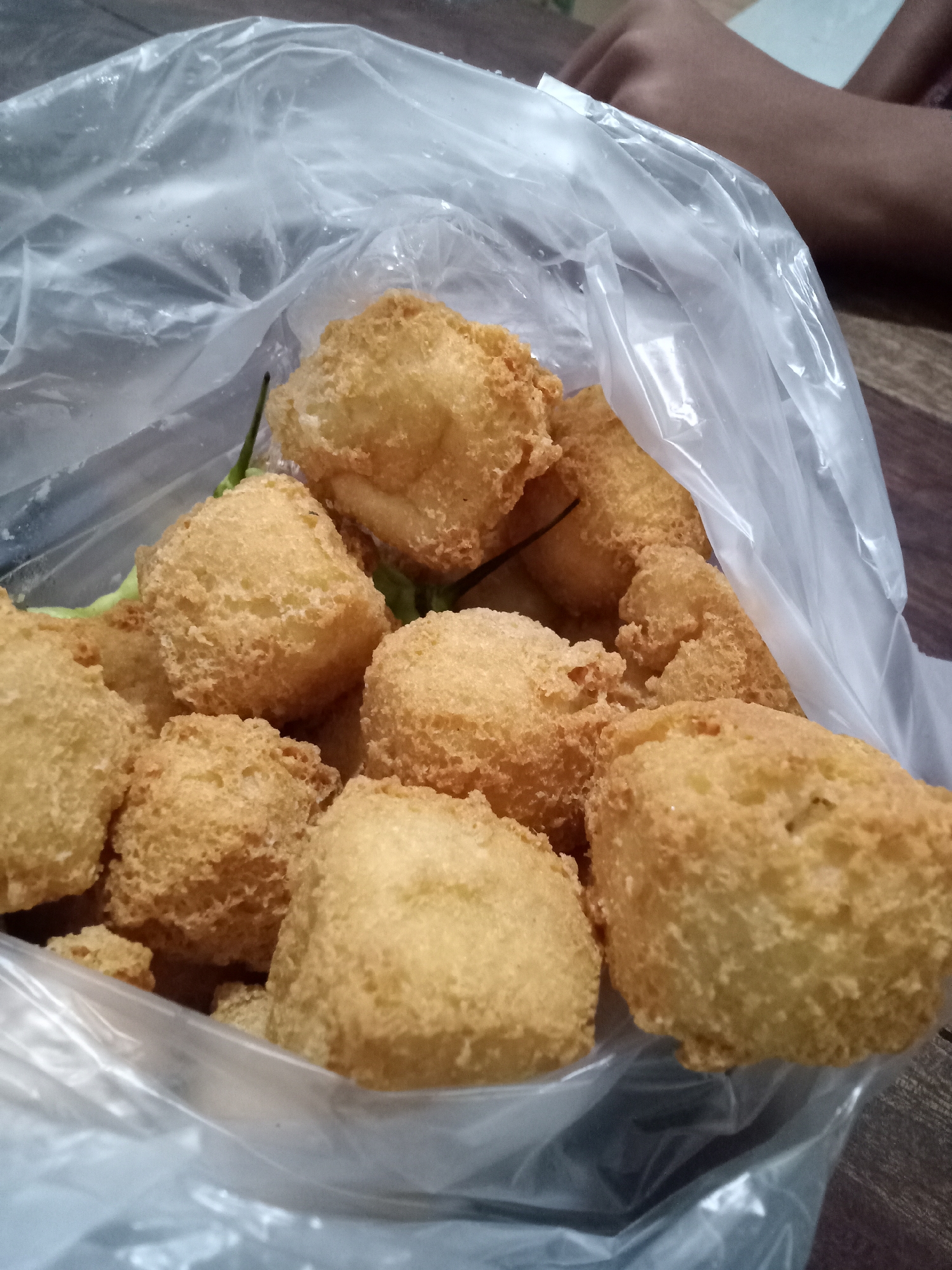 A plastic bag filled with fried tofu, or "tahu goreng," is displayed. The tofu pieces are golden brown and have a fluffy, slightly irregular texture from the batter. Some green chilies are visible interspersed with the tofu. The bag is translucent, allowing the texture and color of the tofu to be clearly seen. The background consists of a wooden surface, suggesting the food is on a table or counter. A portion of a person's arm and hand is visible in the upper right corner, indicating someone is holding or near the bag. The overall impression is of a common street food or home-cooked snack, potentially in Simo, Indonesia, given the context provided, although the image itself does not contain explicit markers of its location.