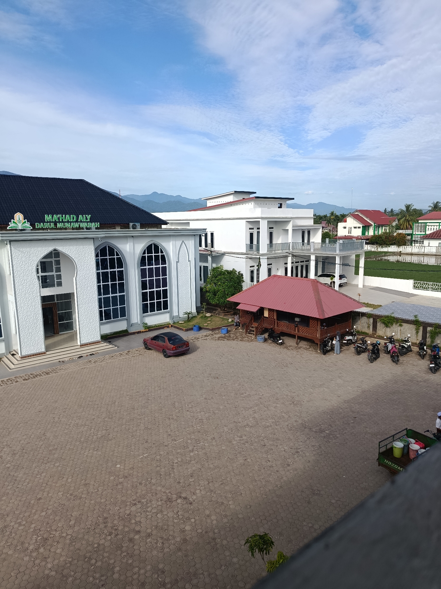 A wide shot captures an outdoor scene at the Ma'had Aly Darul Munawwarah in Peulakancibrek, Indonesia. The foreground is dominated by a light-colored hexagonal paver courtyard. To the left, a white building with arched windows and ornate decorative panels features the school's name prominently displayed in green lettering above the entrance. A dark red sedan is parked in front of this building. In the mid-ground, a reddish-brown wooden building with a red-tiled roof stands on stilts, housing several people and motorcycles. Behind this, a multi-story white building with balconies and dark-trimmed windows is visible. The background shows a lush green landscape with palm trees and mountains under a partially cloudy blue sky. Several motorcycles are parked near the wooden structure, and a small green cart with buckets is positioned in the lower right corner.