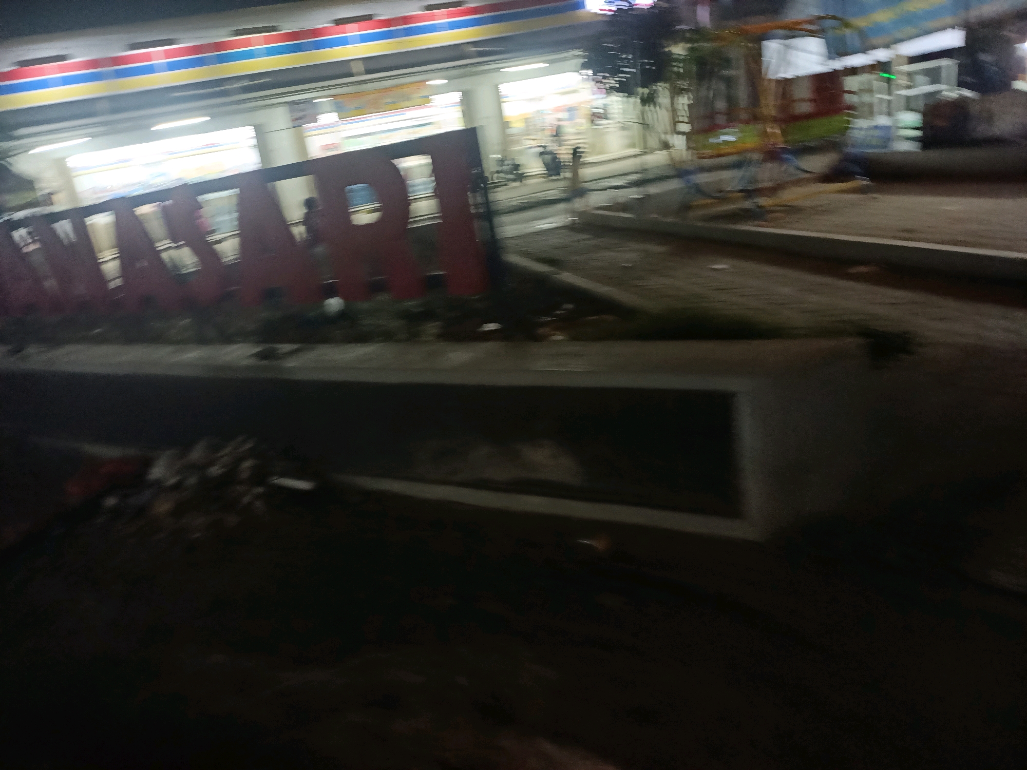 The image depicts a dimly lit street scene at night in Wanajaya, Indonesia, likely in front of a convenience store with a prominent red sign featuring white letters that read "INDOMARET". The store appears to have brightly lit interiors with shelves stocked with goods visible through the windows. In front of the store, a large, three-dimensional red sign with white lettering, partially obscured and out of focus, spells out "INDOMARET". The foreground shows a dark, out-of-focus area with some debris. The background reveals the store's facade with horizontal stripes of red, blue, and yellow above the signage, indicative of a common chain store branding. A portion of what appears to be a playground structure or an amusement ride with colorful elements is visible to the right of the store. The overall impression is one of a commercial area at night, captured with motion blur, suggesting the photo was taken from a moving vehicle.
