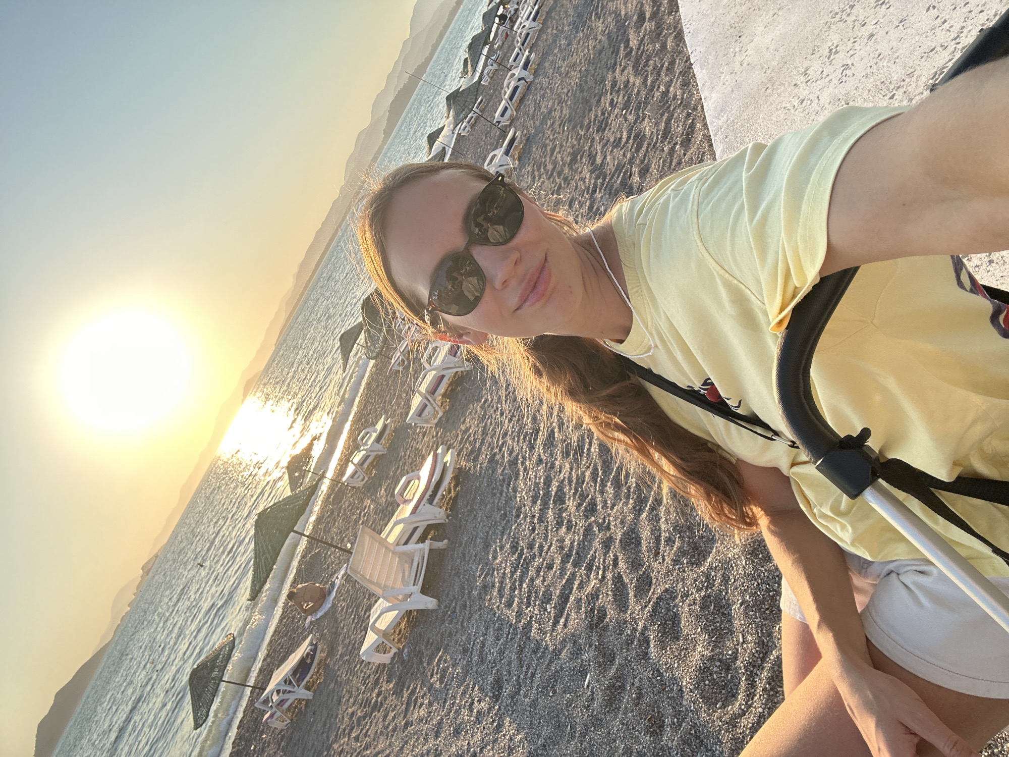 A woman wearing sunglasses and a light yellow t-shirt is taking a selfie on a pebble beach in Fethiye, Turkey. The sun is setting, casting a warm glow over the scene. In the background, lounge chairs and umbrellas line the beach, and the sea stretches out to the horizon, with mountains visible in the distance. The woman is holding a stroller handle, suggesting she may be with a child.