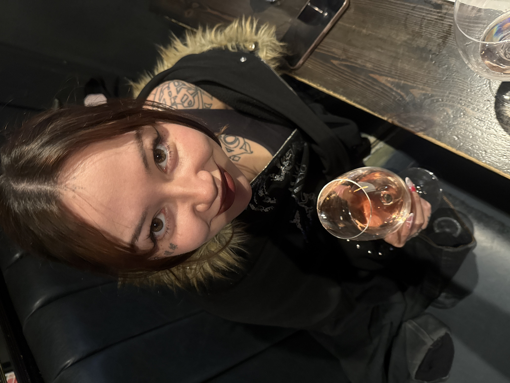 A young woman is captured from a high, slightly tilted angle, looking up at the camera with a playful smile or smirk. She has dark brown hair with bangs, styled over her forehead, and prominent brown eyes. Her face features a small tattoo beneath her left eye. She is wearing a dark jacket with a fur-trimmed hood over a dark top, which also appears to have intricate, possibly tattooed, designs on her arms and shoulder area. Her nails are long and painted a light color.
In her right hand, she holds a stemmed glass containing a rose-colored beverage, which appears to be wine or a cocktail. The setting is an indoor establishment, characterized by a dark wooden table in front of her. To her right, on the table, a smartphone and another partially visible glass are present. She is seated on dark, padded, booth-style seating. The lighting is relatively dim, suggesting an evening or night-time ambiance in a bar, restaurant, or lounge. No discernible text is visible on any objects or the person's tattoos.