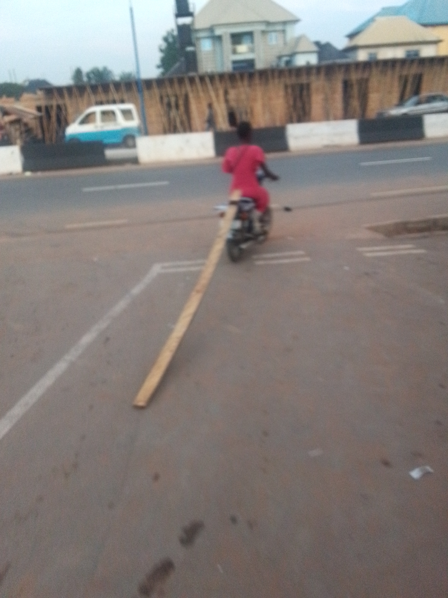 A person on a moped is dragging a long piece of lumber on a paved road in Unknown, Unknown. The person is wearing a red outfit. Behind the person are several buildings and a vehicle.