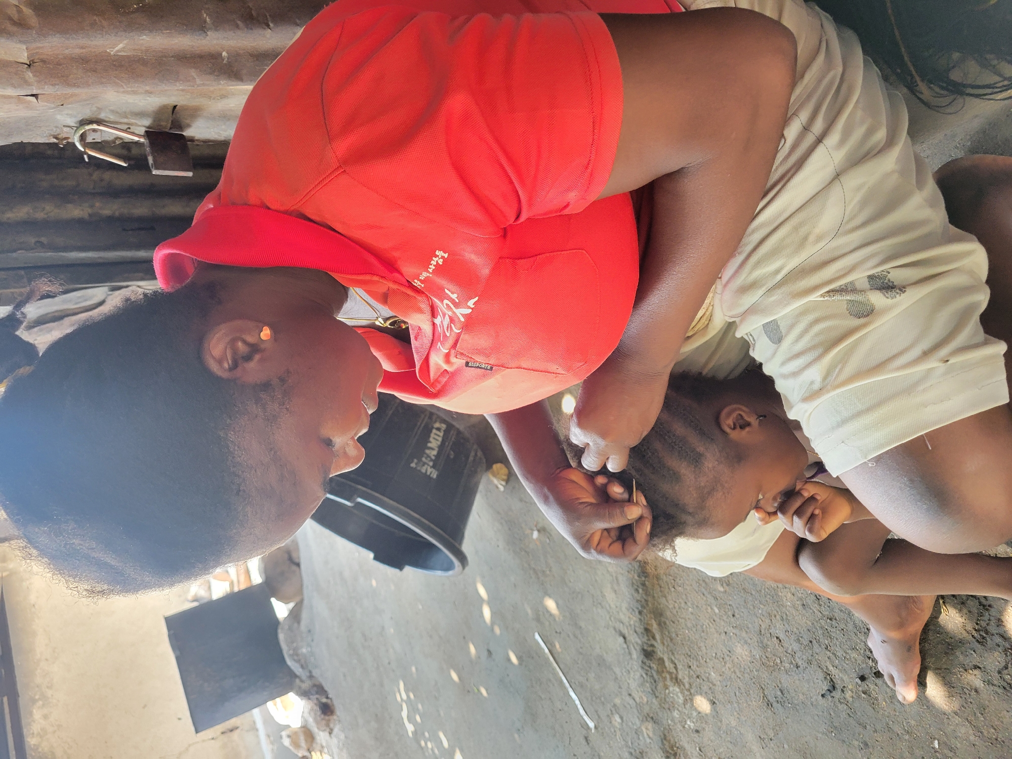 An adult is braiding a child's hair in Madalla, Nigeria. The adult, wearing a red top, is using a tool, possibly a comb or needle, to style the child's hair into braids. The child, wearing a white shirt, is seated on the ground with their hands near their mouth, possibly in discomfort. A black bucket is near the adult. The ground appears to be rough and uneven. A partial view of a metal structure and debris can be seen in the background.