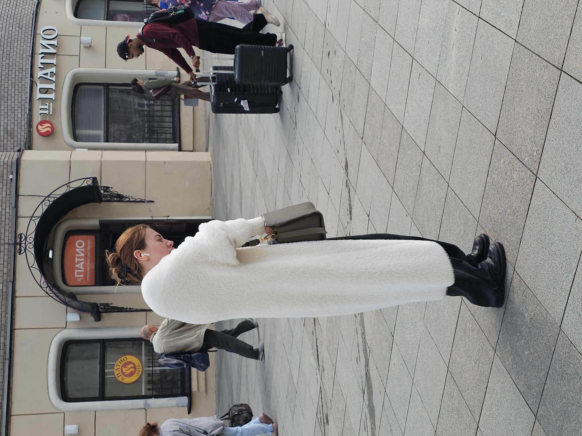 The image depicts a street scene in Moscow, Russia, likely in front of a restaurant or establishment with Cyrillic signage reading "ПАТИО" (Patio). A woman with reddish-brown hair pulled back is the primary subject, facing down and examining something in her hands while wearing a long, fluffy white coat and black boots. She is also carrying a grey handbag. Several other individuals are visible in the background: a man with a beard and a cap is pulling a black rolling suitcase, and another man in a beige jacket with a backpack is walking away from the camera. Further in the background, a person in a patterned outfit is partially visible. The scene takes place on a paved sidewalk composed of square tiles, with a building featuring arched doorways and windows in the background. The lighting suggests it is daytime.