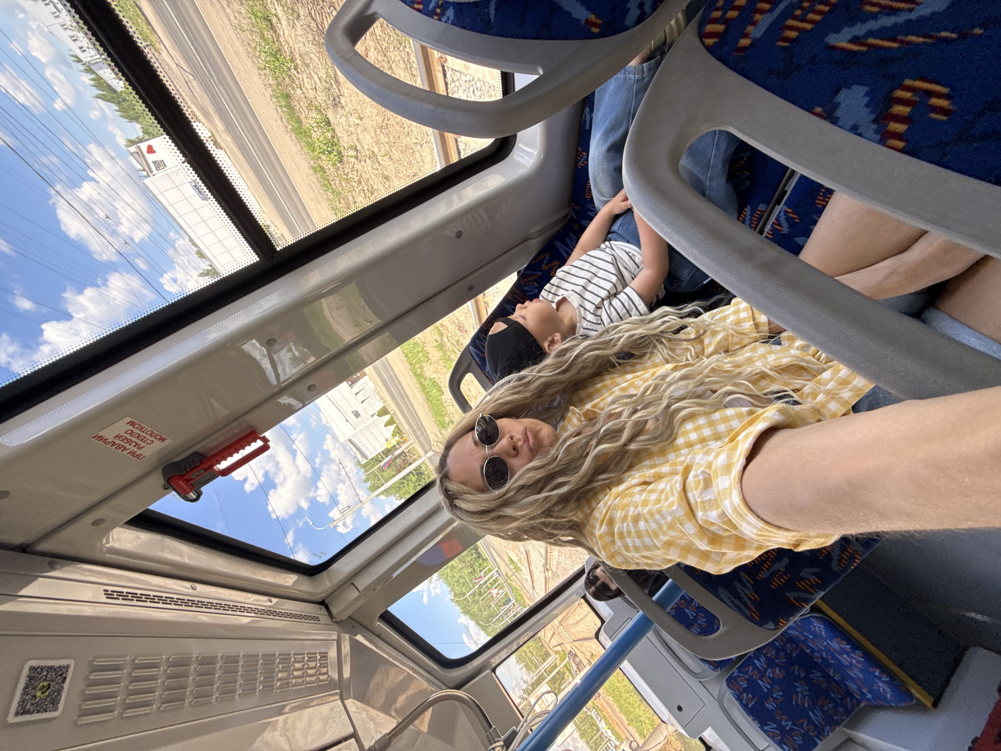 The photo depicts two individuals on a bus in Заволжье, Russia. An adult with blonde, curly hair and sunglasses is visible, wearing a yellow and white checkered shirt. A young child, wearing a black face mask and a white shirt with black stripes, is lying across the seats. The bus's interior features blue and patterned seats, overhead handles, and windows showing the passing landscape, including a road, fields, buildings, and a cloudy sky. Emergency equipment with Russian script is mounted near the windows.