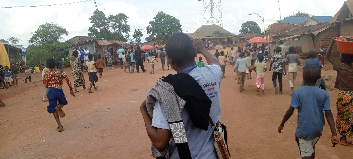 This image presents an expansive outdoor scene on an unpaved, reddish-brown dirt surface, likely a street or open communal area, located in the city of Fizi, Democratic Republic of the Congo. The sky overhead is overcast. Various single-story structures, constructed from brick or corrugated metal, line the perimeter, with scattered green trees visible throughout the background and mid-ground. A prominent metal lattice communication tower stands in the distant right-center.
The scene is populated by a substantial gathering of individuals, predominantly children, with several adults interspersed. In the immediate foreground, an individual with dark skin is shown from the back, wearing a light-colored t-shirt with the partial text "AAIYA" visible on the back. A black garment and a lighter, patterned fabric are draped over their left shoulder, from which a patterned-strap bag also hangs. Their right arm is raised, extended forward.
Numerous children are actively moving across the dirt ground; some are running, others are walking. A child in the mid-left foreground is in motion, wearing a brightly patterned long-sleeved top and blue shorts. In the mid-right, another child carries a red basin balanced on their head. A group of adults, some dressed in white and blue shirts, stands near a structure on the left side of the frame. A red market umbrella is visible in the mid-ground, slightly to the left of the center. Yellow jerrycans are present near a structure on the far left.