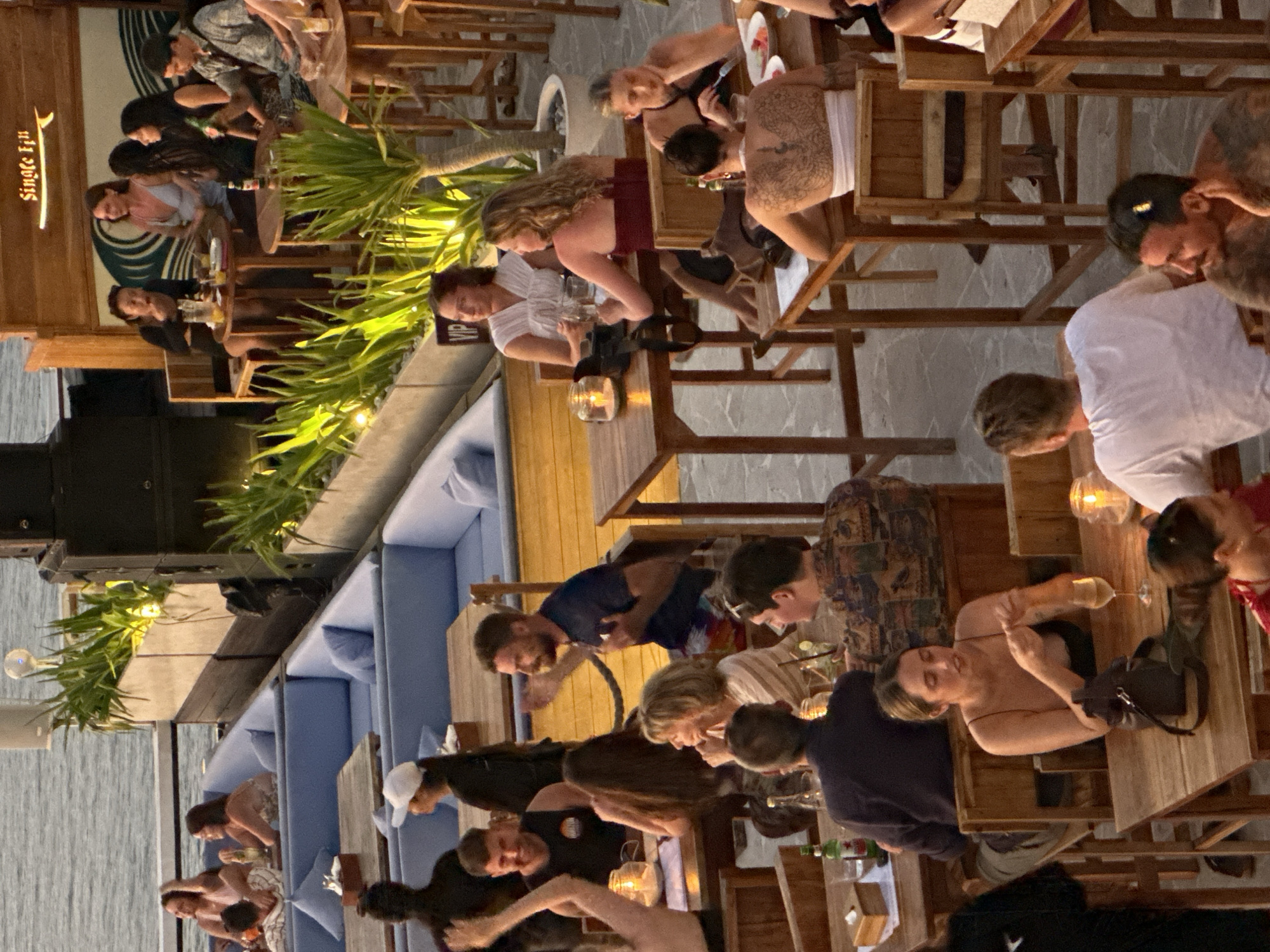 The image shows an outdoor restaurant or bar setting in Kuta Selatan, Indonesia. It is populated with numerous people who appear to be socializing, eating, and drinking at various tables. Most are seated on wooden chairs, and the tables are set with glassware, some with candles. The restaurant appears to be situated near a waterfront, potentially overlooking the ocean. Some blue seating arrangements (likely lounge seating) are visible in the background. Palm trees and other greenery are dispersed throughout the space, adding to the ambiance. A sign "Single Fin" is visible, potentially identifying the venue.