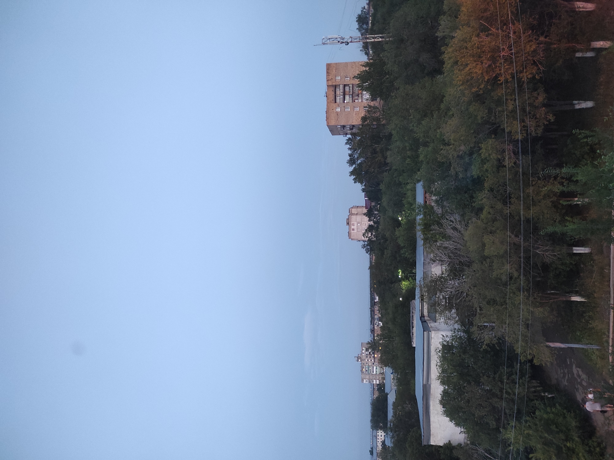 An aerial view captures a city skyline under a pale blue sky at dusk. In the foreground, a dense canopy of dark green trees obscures the lower half of the image. Two figures, a man and a child, are visible walking on a dirt path amidst the foliage. A low-rise building with a blue roof is partially visible above the trees. Further back, several multi-story apartment buildings rise above the treeline. One prominent brick apartment building stands to the right of center, featuring numerous balconies. A cellular tower with multiple antennas is situated to the right of this building, its structure illuminated by the fading light. In the distance, more apartment blocks and smaller structures are scattered across the horizon, indicating an urban environment. The overall scene suggests a residential area in Shakhtinsk, Kazakhstan, during the transitional period between day and night.
